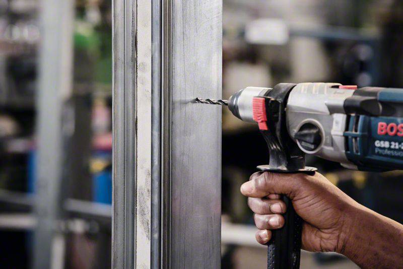 Osoba wierca wiertarką w metalowy materiał. Otoczenie ma charakter przemysłowy.