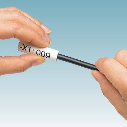 Dwie ręce trzymają czarny kabel z białą etykietą, na której widnieje napis '-X1:009'. Tło jest niebieskie.