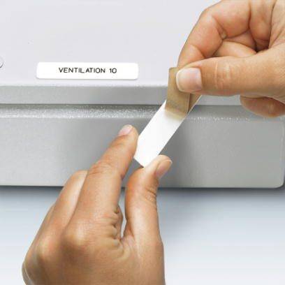 Jedna osoba przykleja brązowy pasek z napisem 'Wentylacja 10' do szarego pudełka.