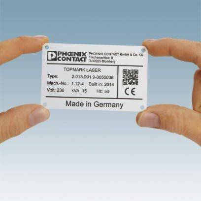 Dwie ręce trzymają metalową etykietę z napisem 'Made in Germany'. Producent: Phoenix Contact. Typ: Topmark Laser.
