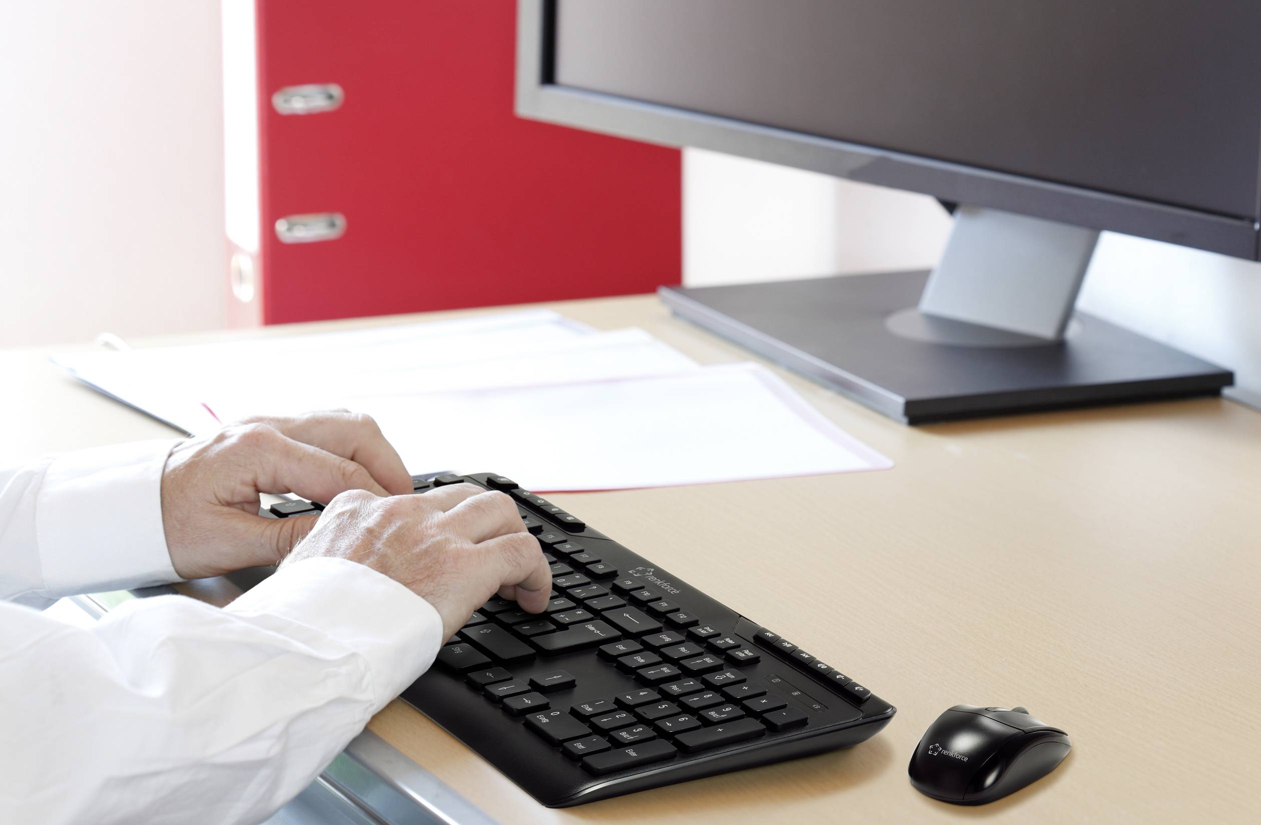Osoba pisze na klawiaturze komputera przed ekranem, z czerwoną teczką w tle na biurku.