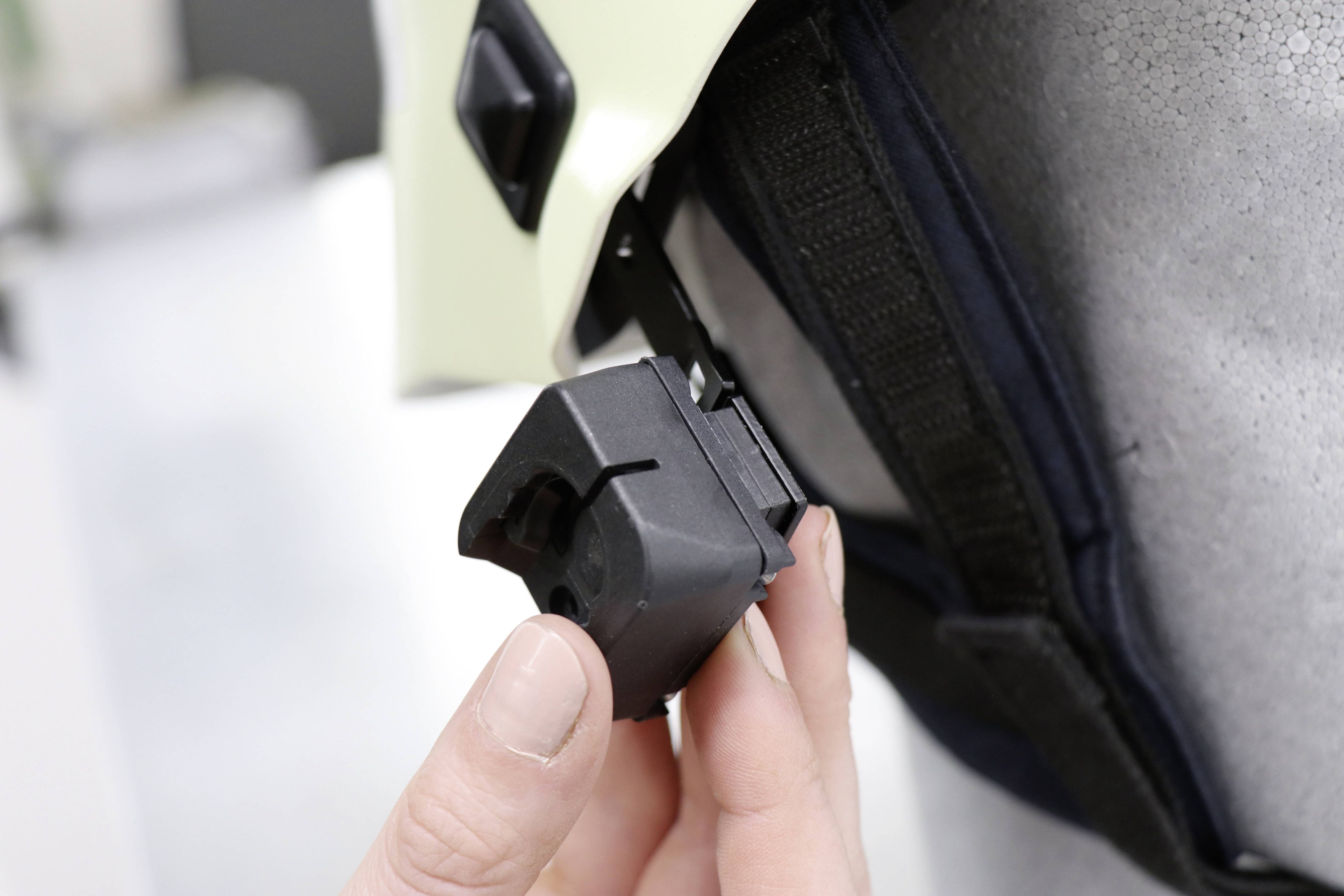 Osoba trzyma czarny, sześcienny przyrząd zamocowany z boku kasku. Wygląda na to, że jest to urządzenie zabezpieczające.