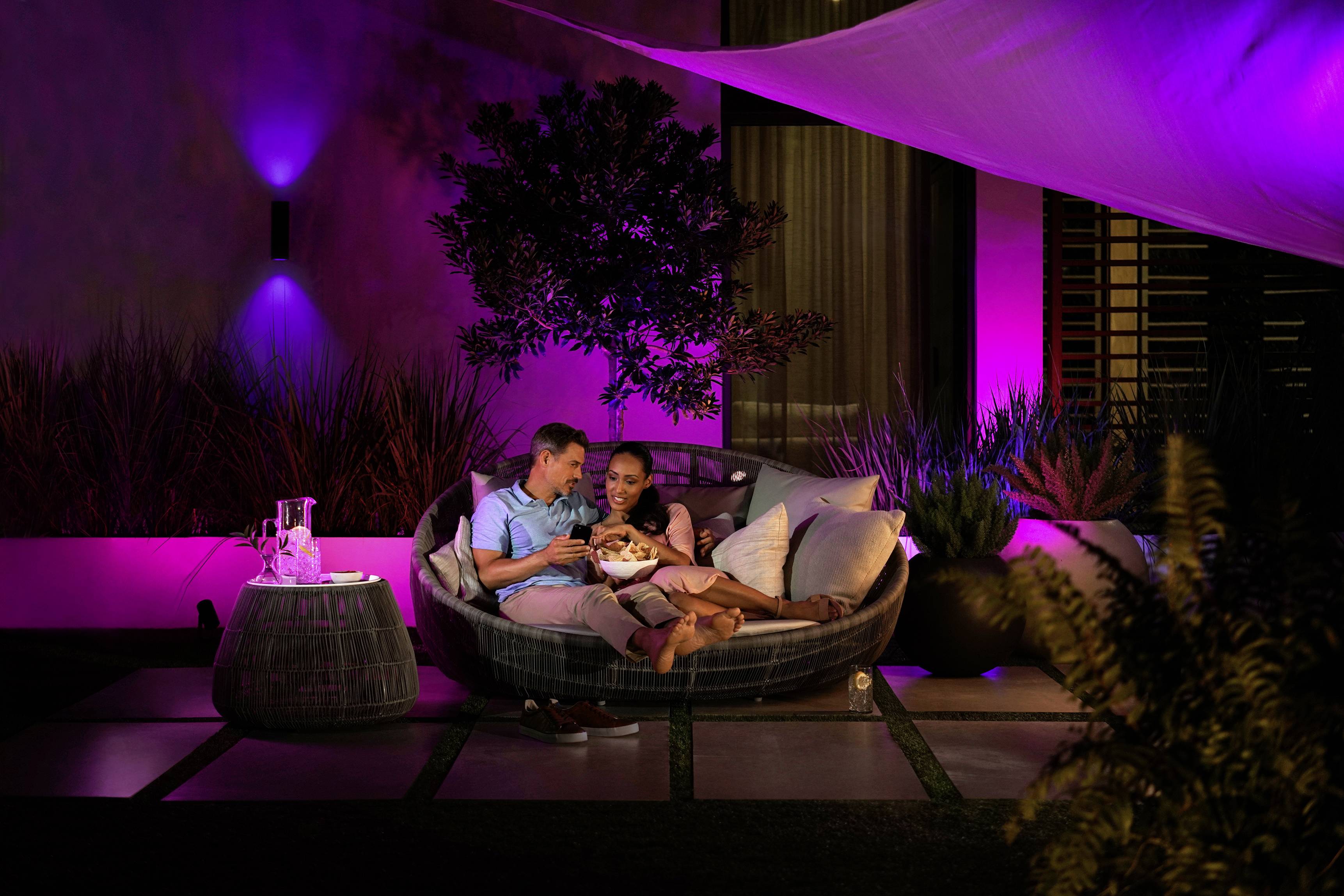 Para siedzi wieczorem spokojnie w oświetlonej przestrzeni outdoorowej pod zadaszeniem, otoczona fioletowym światłem.