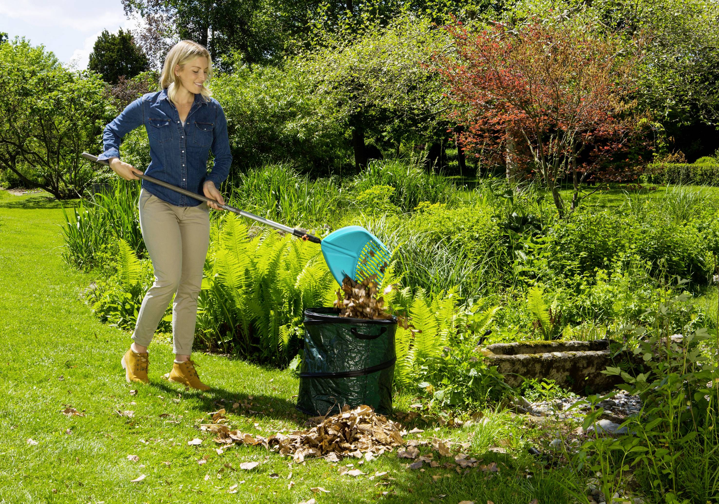 Kobieta ubrana w strój casualowy grabia jesienne liście w zielonym ogrodzie i zbiera je do kosza na śmieci. W tle widoczne drzewa i krzewy.