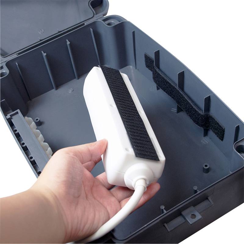 Dłoń trzyma podłużne, białe urządzenie w otwartej szarej obudowie. Urządzenie posiada paski rzepowe.