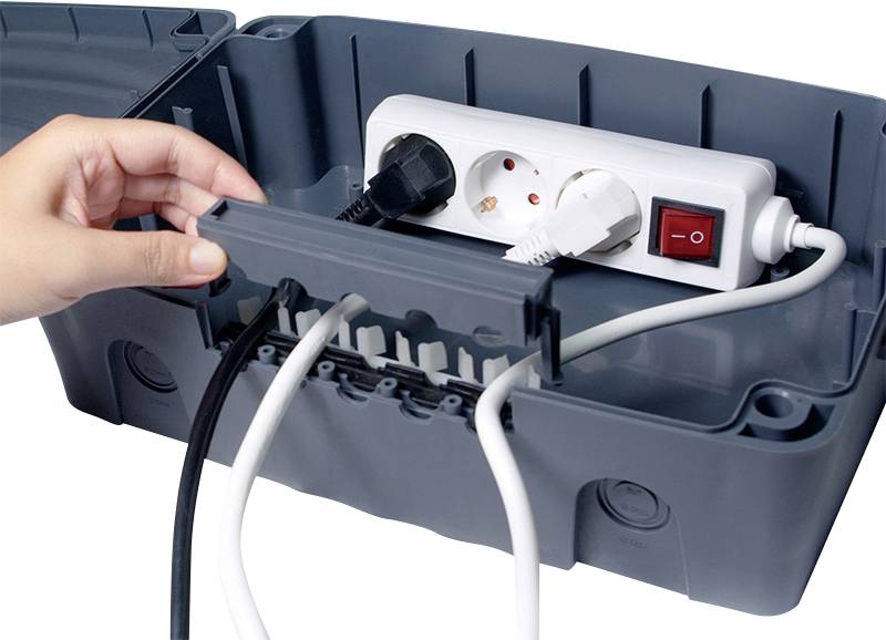 Ręka otwiera szare plastikowe pudełko z białym przedłużaczem i kilkoma wetknętymi czarnymi i białymi kablami.