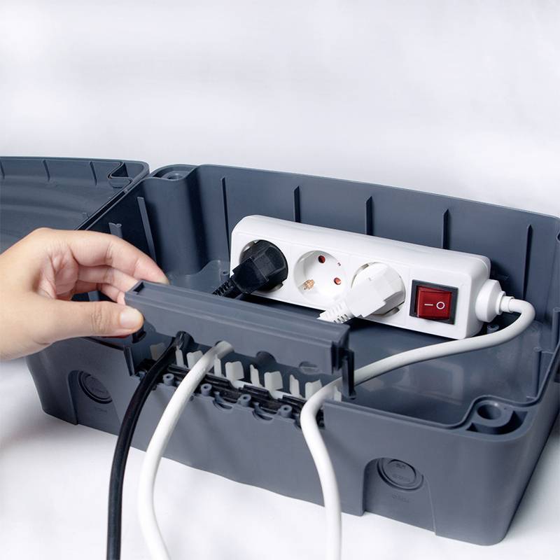 Dłoń otwiera skrzynkę kablową, w której znajduje się rozdzielacz elektryczny z podłączonymi przewodami i włącznikiem sieciowym.