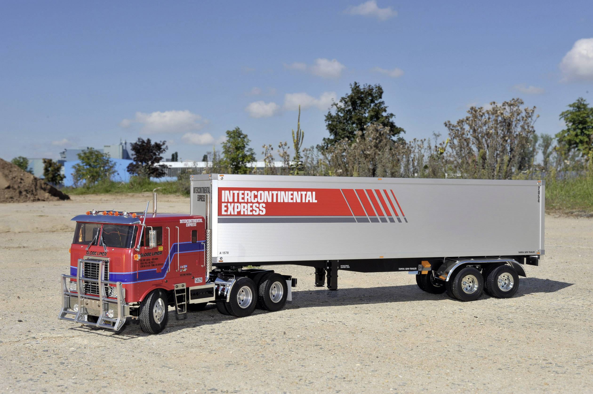 Czerwona ciężarówka siodłowa z napisem 'Intercontinental Express' na srebrnej przyczepie stoi na żwirowym placu.