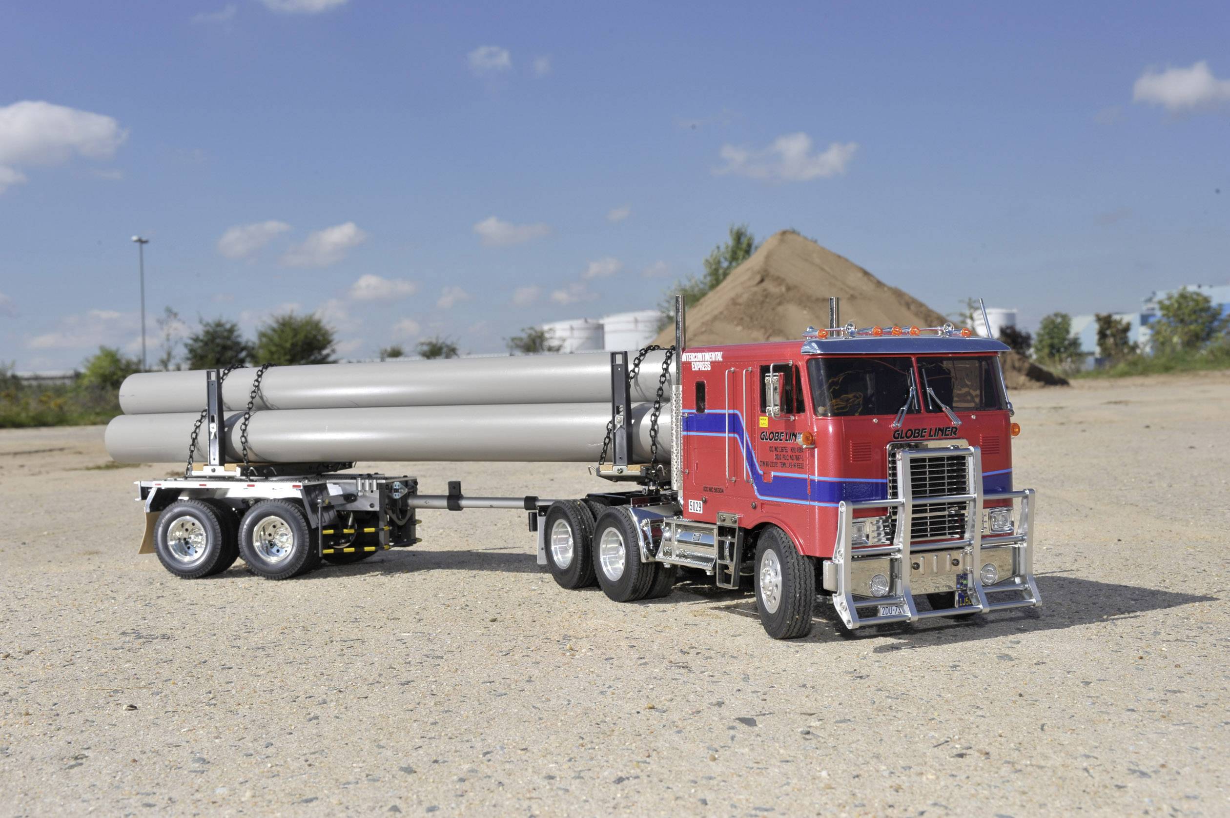 Czerwona ciężarówka siodłowa z niebieskim paskiem holuje przyczepę z długimi rurami na żwirowym placu pod błękitnym niebem.