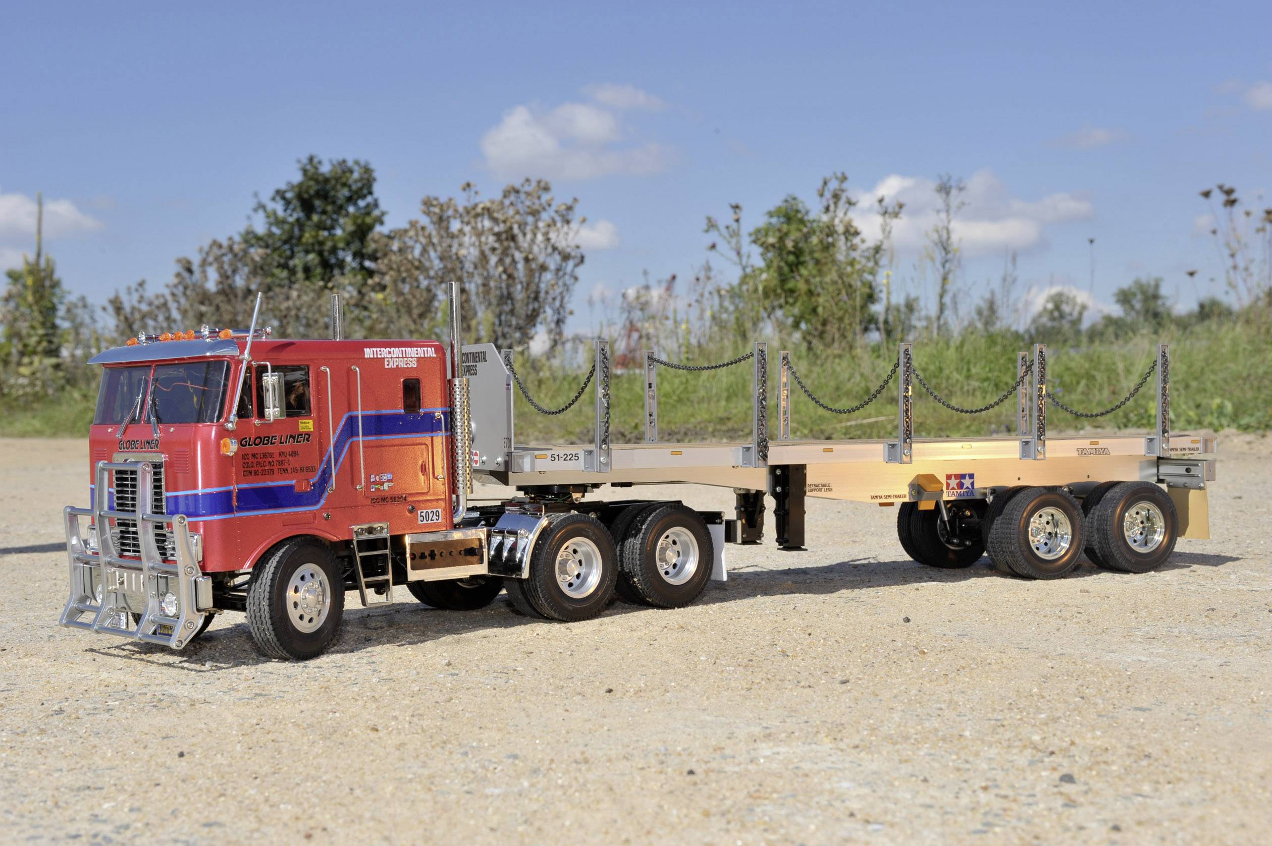 Czerwona ciężarówka z otwartą przyczepą stoi na piaszczystym placu, otoczona drzewami i niebieskim niebem w tle.