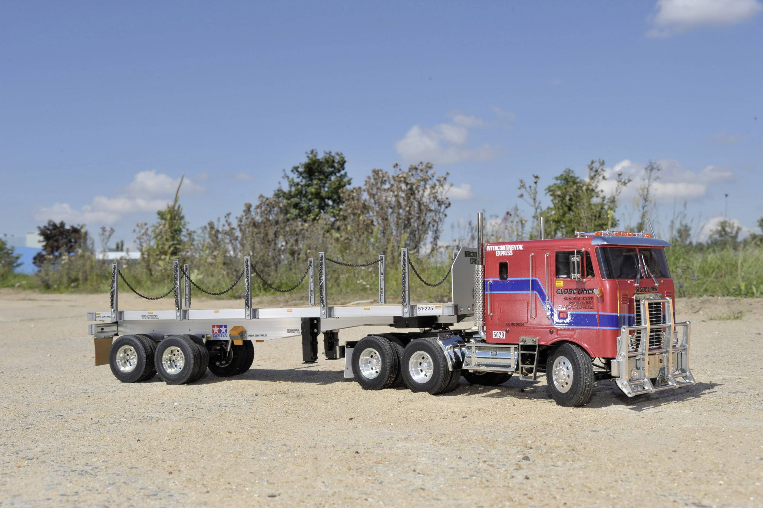 Ciężarówka z czerwoną kabiną i płaską przyczepą bez ładunku, stojąca na piaszczystym terenie z drzewami w tle. Czysty, jasny niebo.
