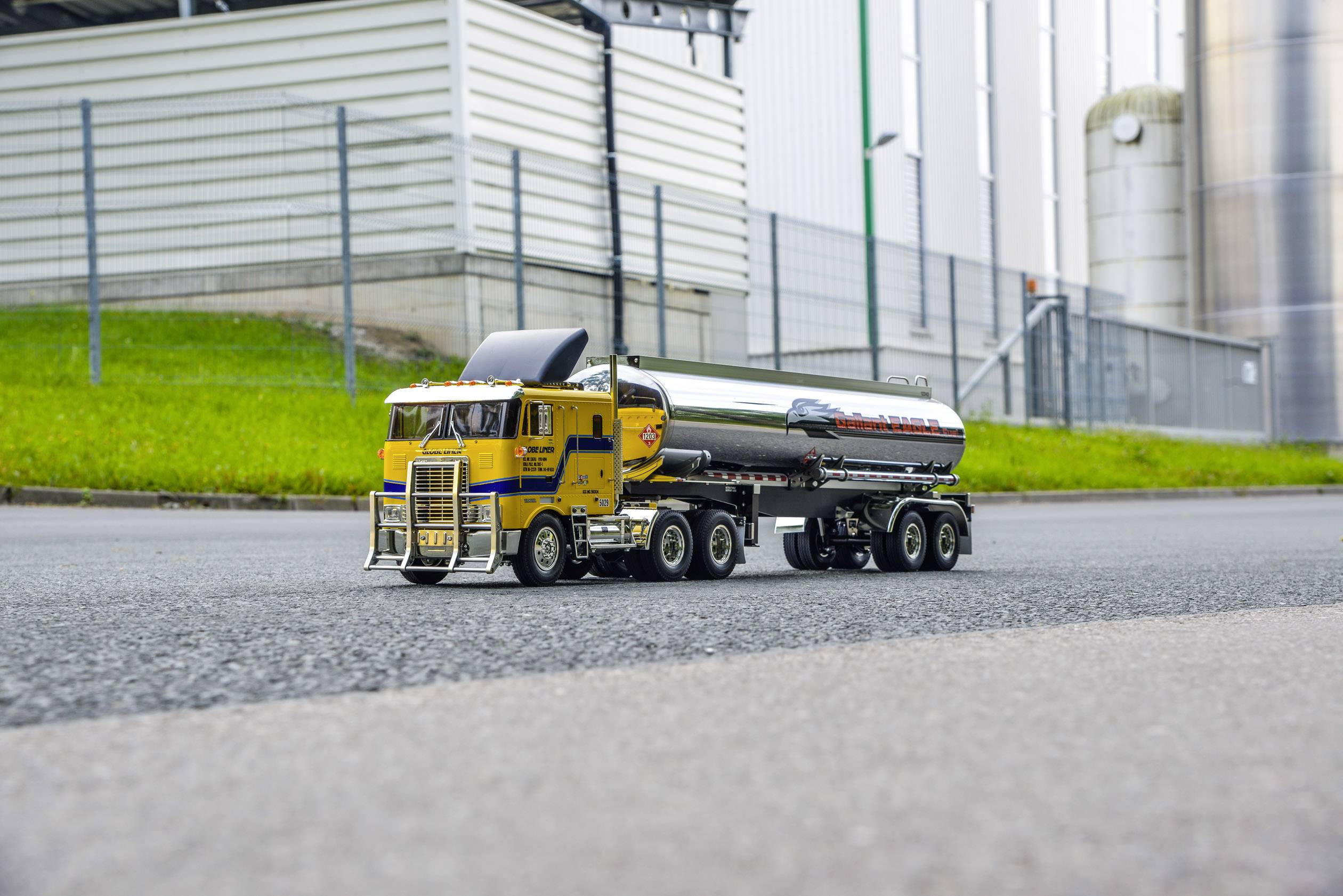 Żółta modelowa ciężarówka z przyczepą cysterną stoi na drodze przed zielonym trawnikiem i przemysłowym otoczeniem w tle.