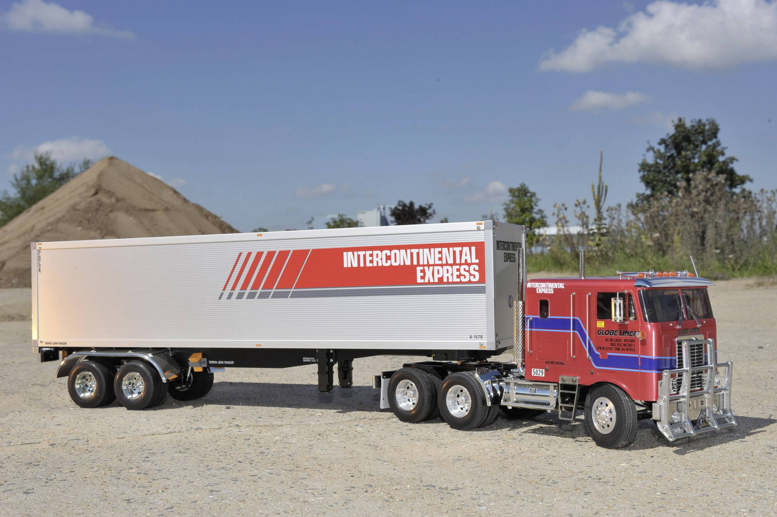 Ciężarówka z przyczepą, na której widnieje napis 'Intercontinental Express', stoi na placu budowy, w tle widać stosy piasku i drzewa.