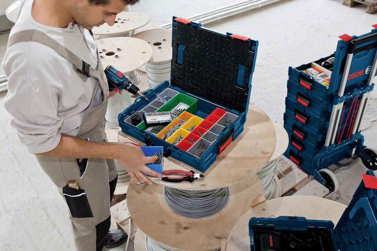 Rzemieślnik segreguje narzędzia w otwartej skrzynce narzędziowej, która stoi na rolce kabla. Obok niego leży stos kolejnych skrzynek.