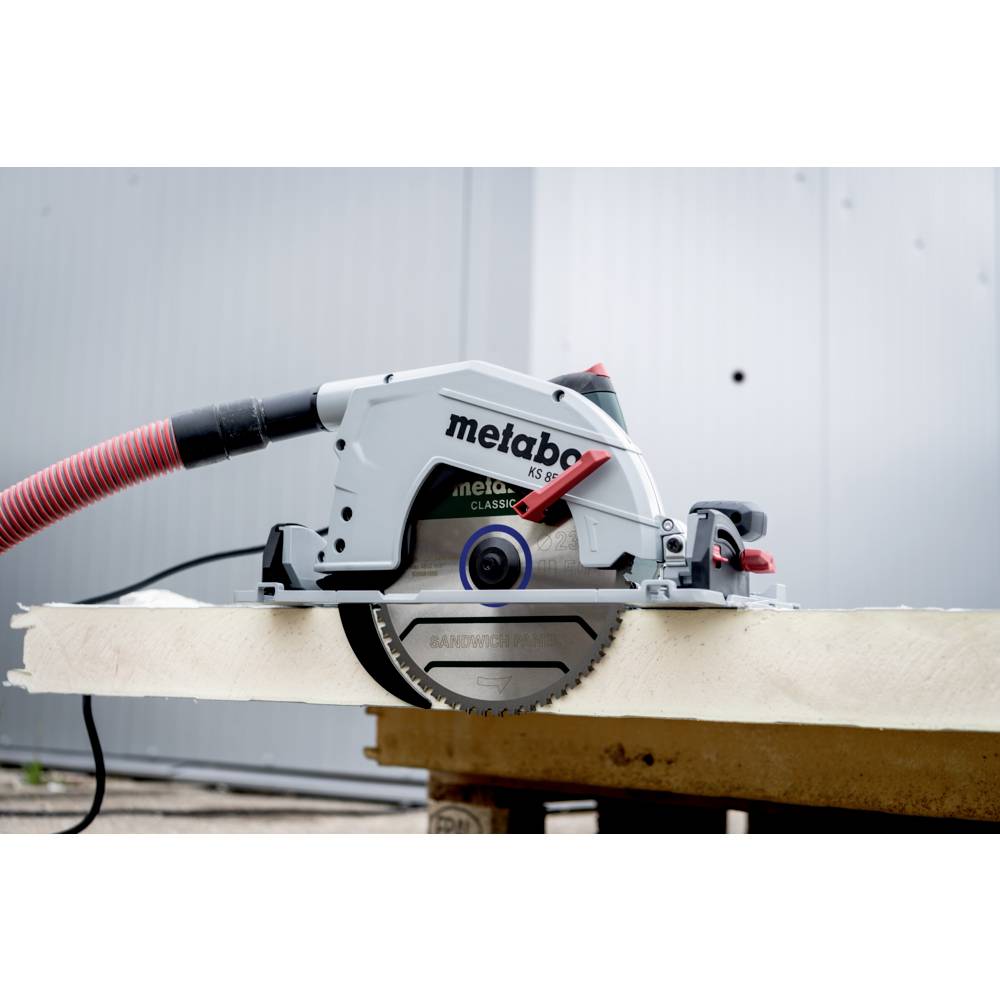 Piła tarczowa spoczywająca na drewnianej belce z czerwonym wężem, używana do cięcia drewna. W tle rozmyte przemysłowe środowisko.