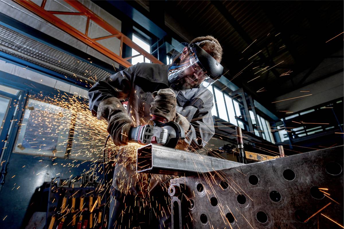 Robotnik w ochronnych okularach i kasku szlifuje kawałek metalu w warsztacie. Od szlifierki odpryskują iskry.