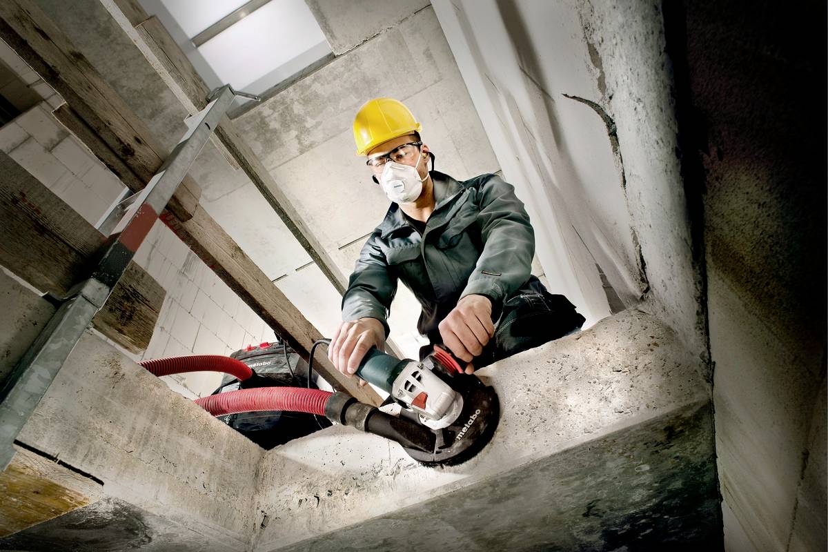 Pracownik w okularach ochronnych, kasku i masce przeciwpyłowej używa szlifierki kątowej przy betonowej ścianie.