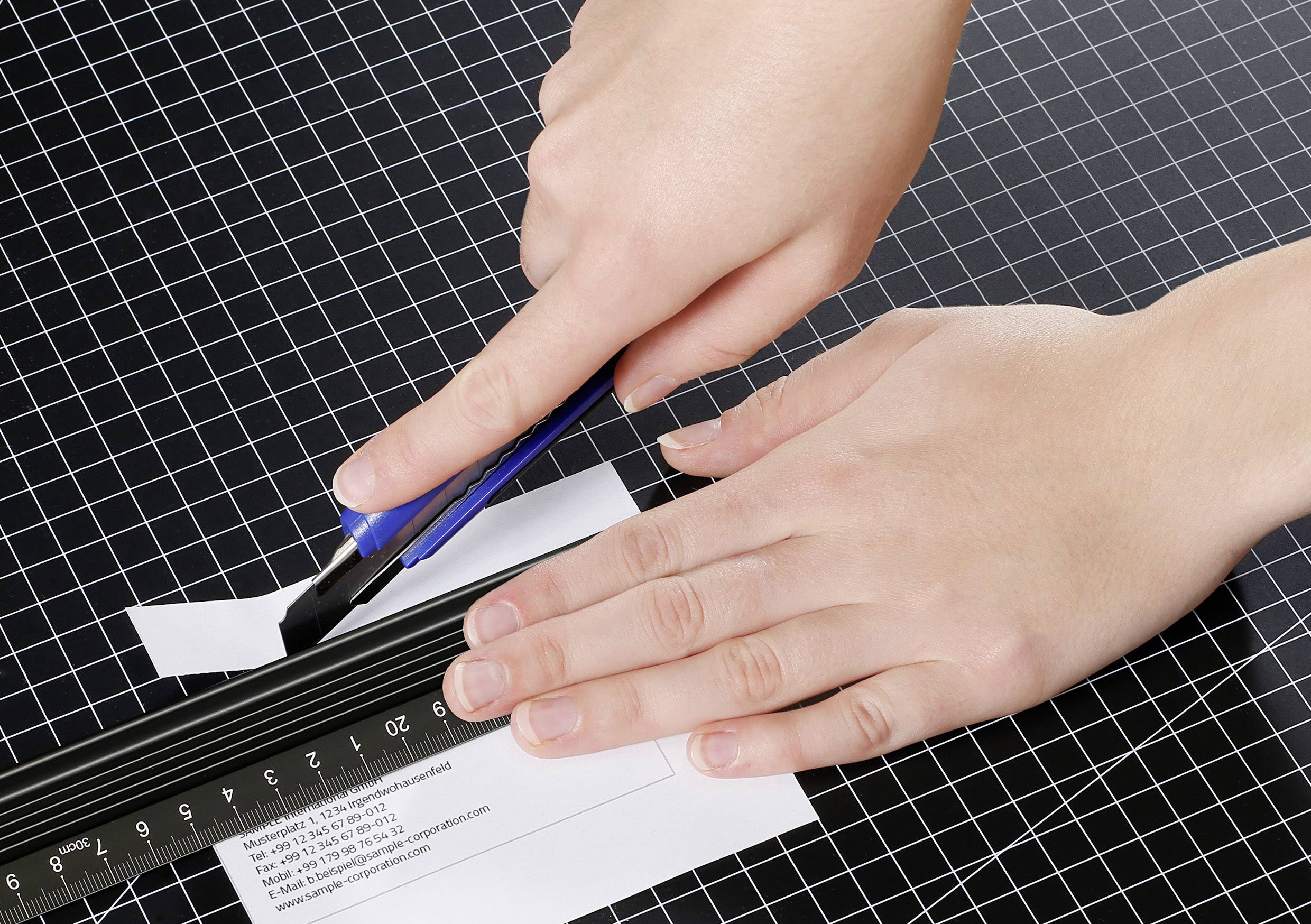 Osoba używa noża do dywanów i linijki, aby ciąć papier na stole w kratkę.