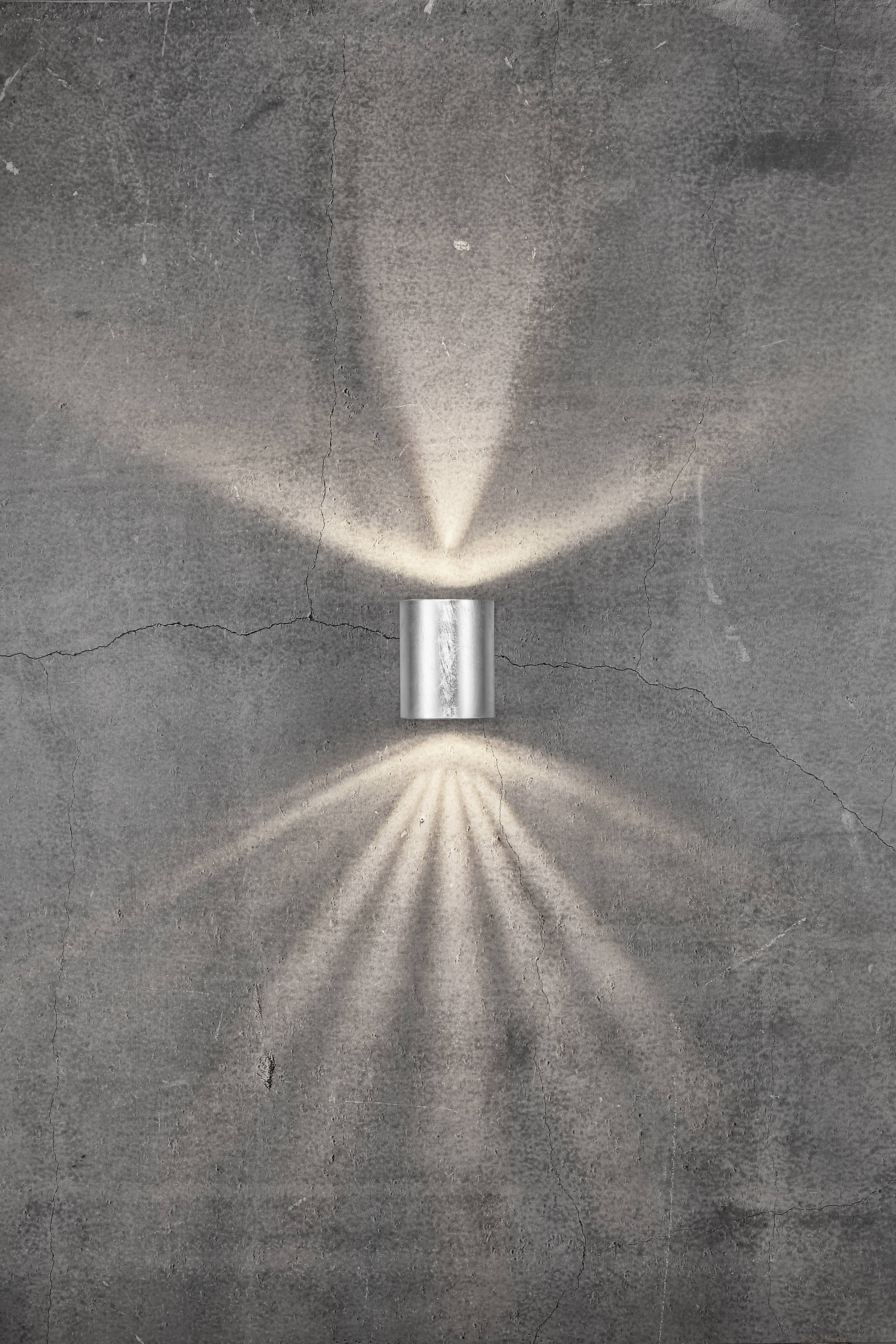 Kinkiet świeci w nowoczesnym designie na szarej betonowej ścianie, tworząc promieniujący wzór świetlny skierowany w górę i w dół.