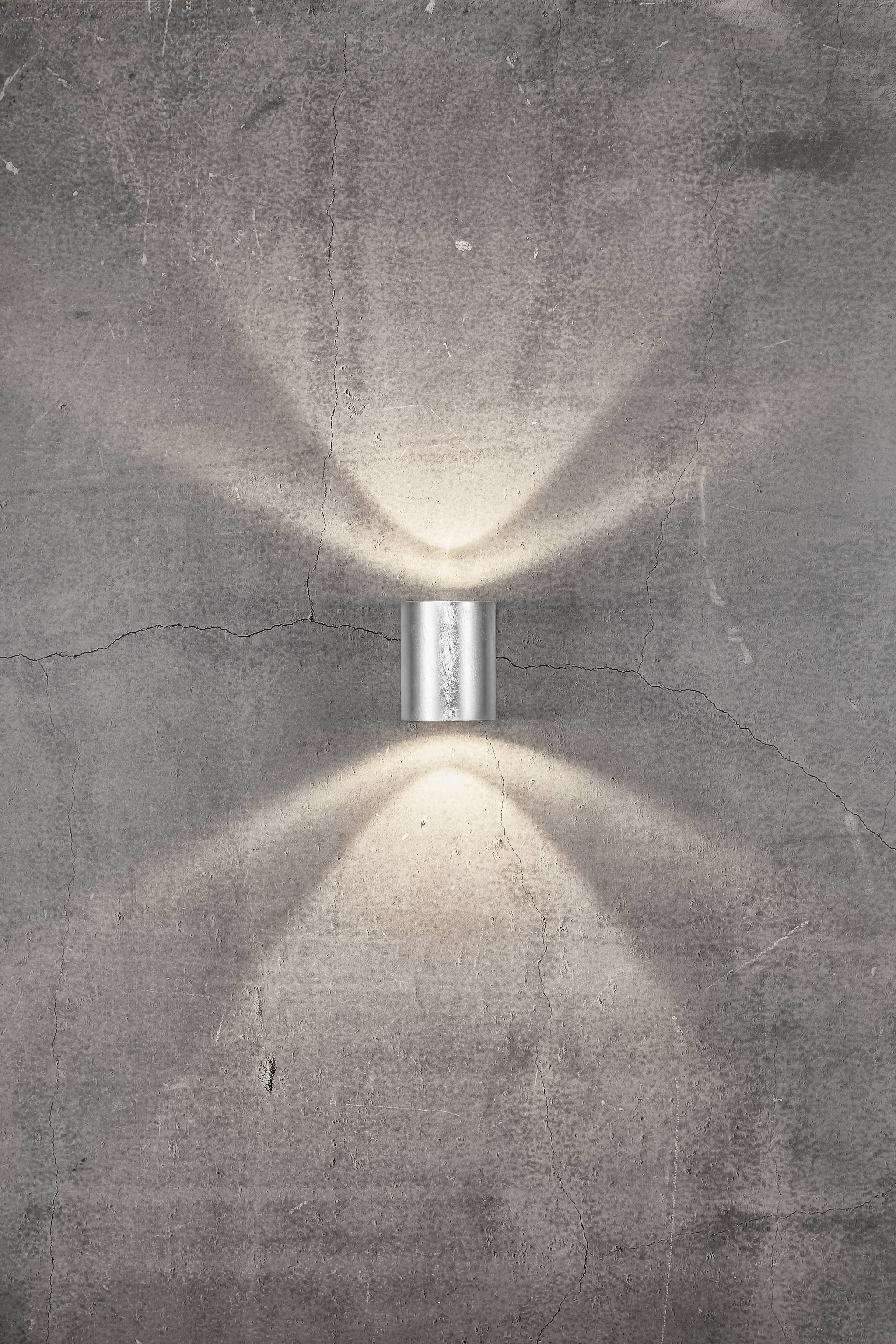 Cylindryczna lampa ścienna, która emituje światło zarówno w górę, jak i w dół, zamocowana na chropowatej ścianie betonowej, tworząc symetryczne wzory świetlne.