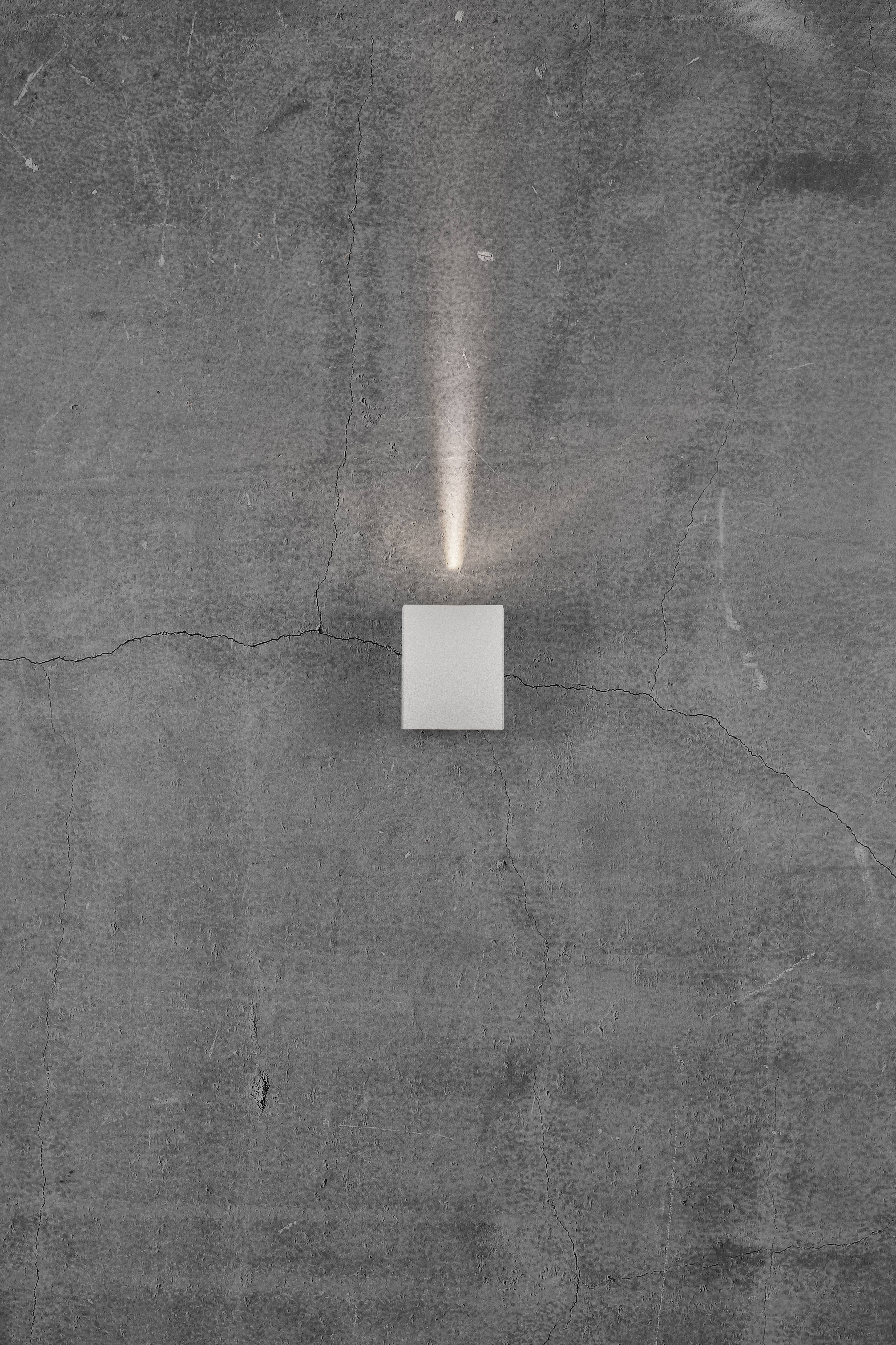 Mały, prostokątny, biały kinkiet świeci w górę i w dół na szarą, lekko spękaną ścianę betonową.