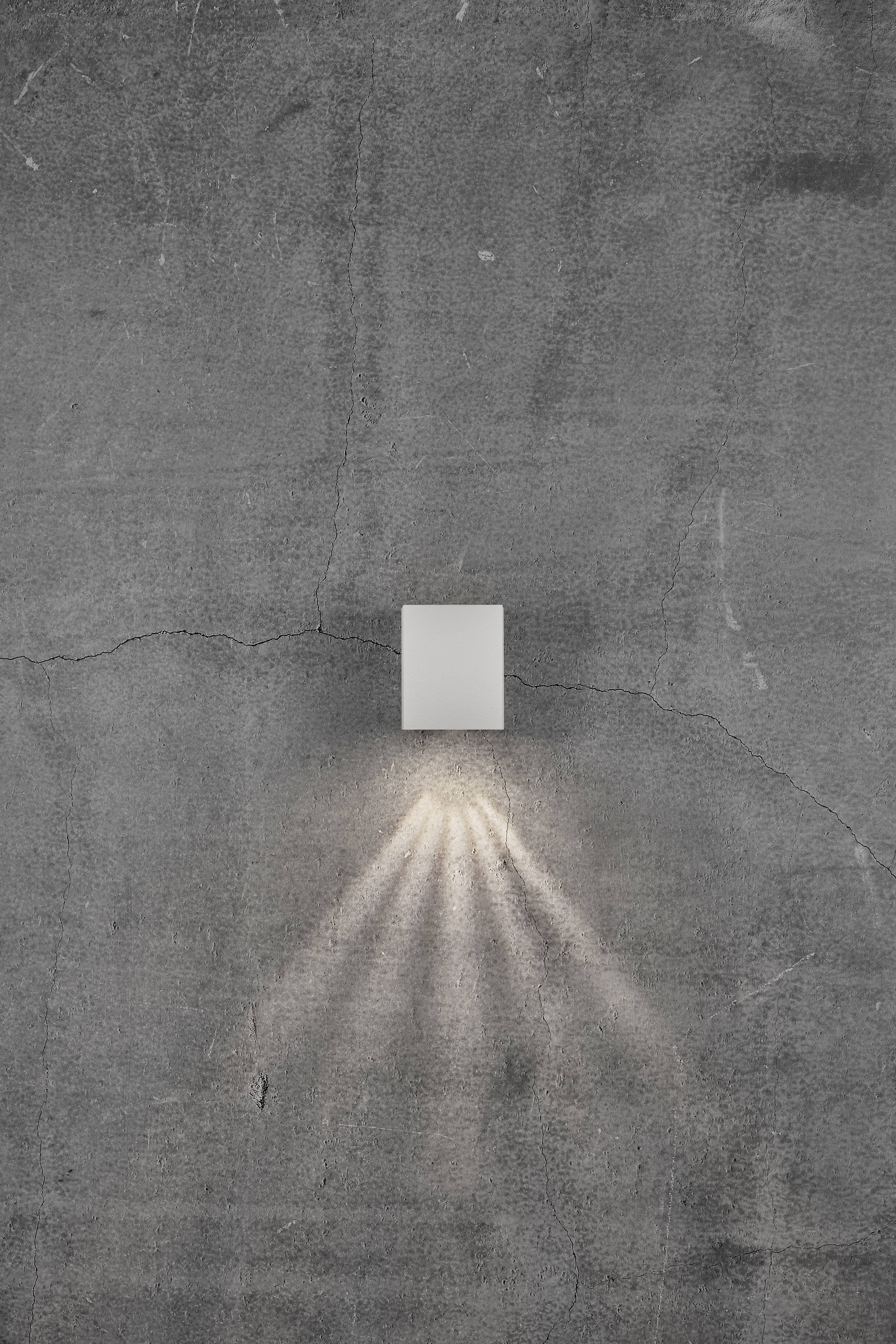 Kinkiet na szarej ścianie betonowej, rozpraszający światło promieniście w dół.