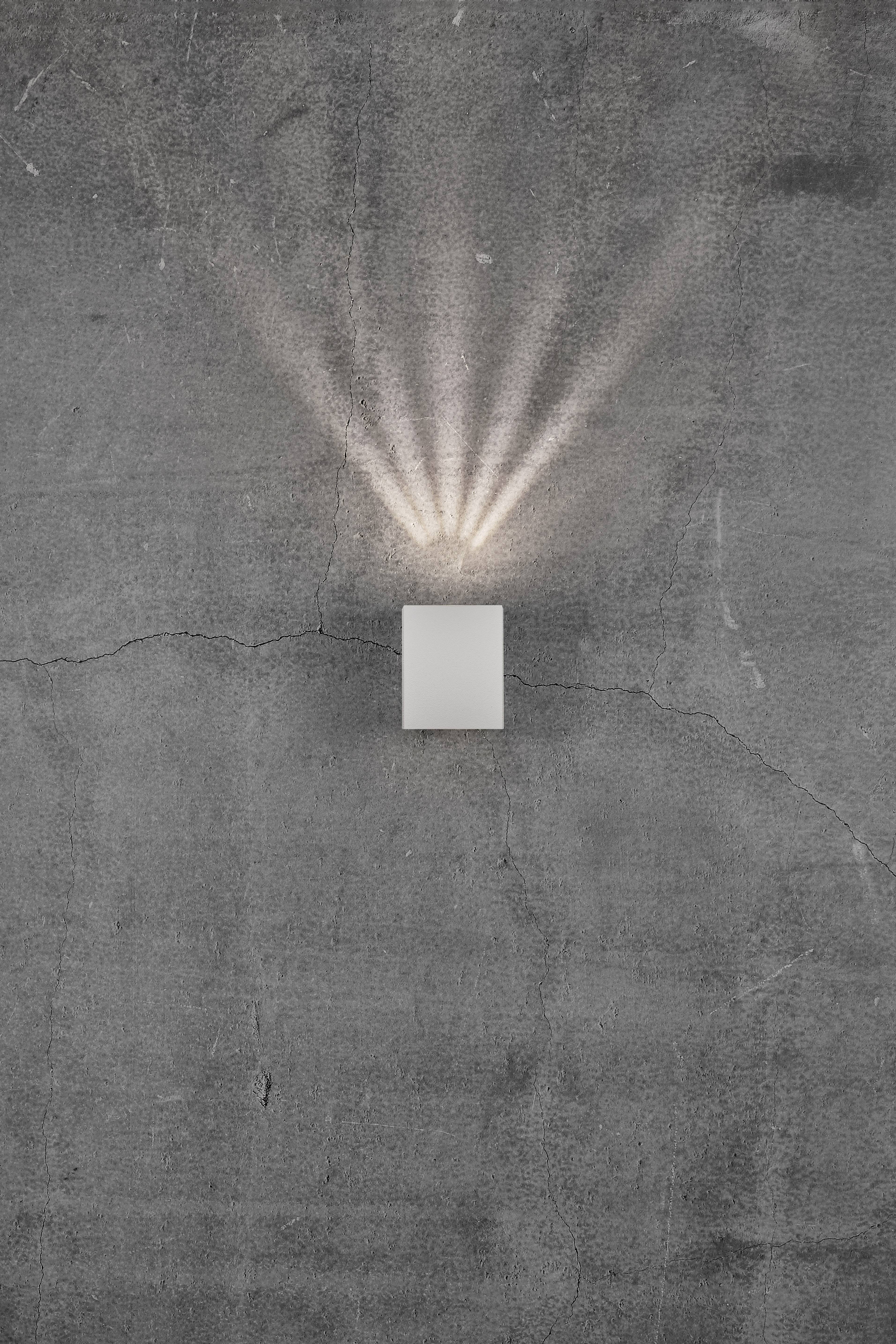 Kinkiet na szarej ścianie betonowej, rzucający smugi światła w górę i na boki, tworzący dekoracyjny wzór efektów świetlnych.