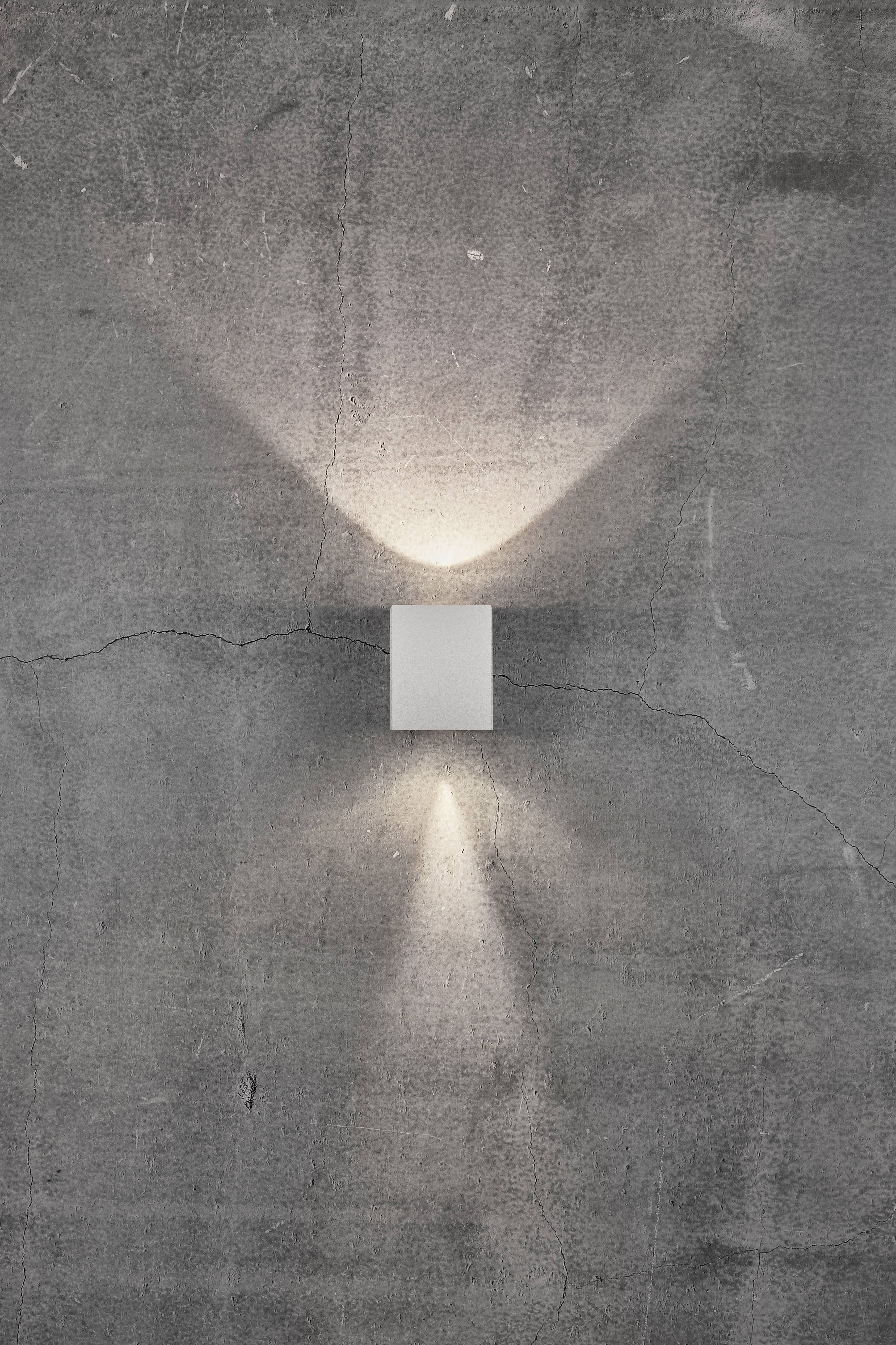 Kinkiet na szarej ścianie betonowej, który rzuca światło w górę i w dół, tworząc geometryczny wzór świetlny.
