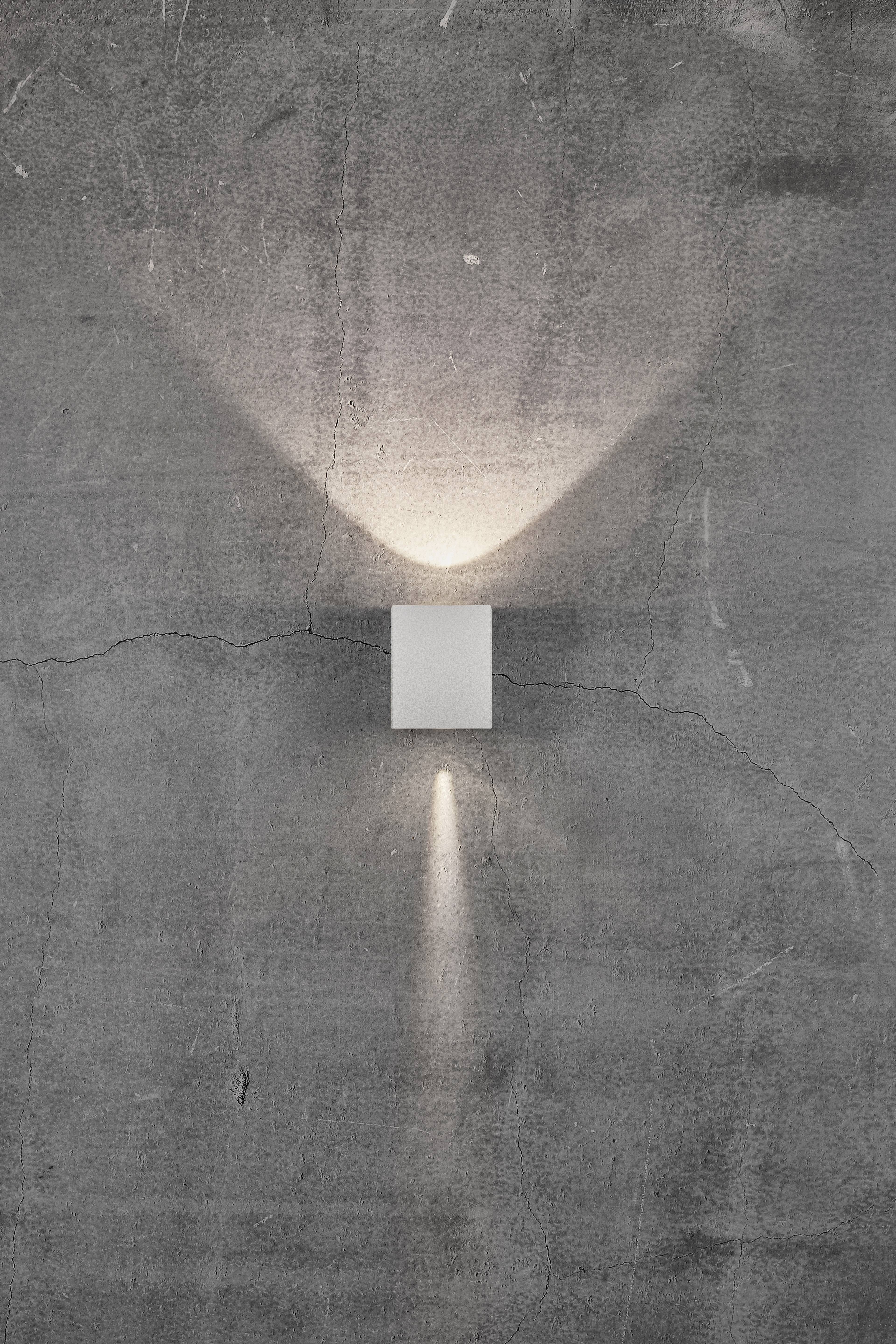 Kinkiet na szarej ścianie, emitujący światło w górę i w dół.