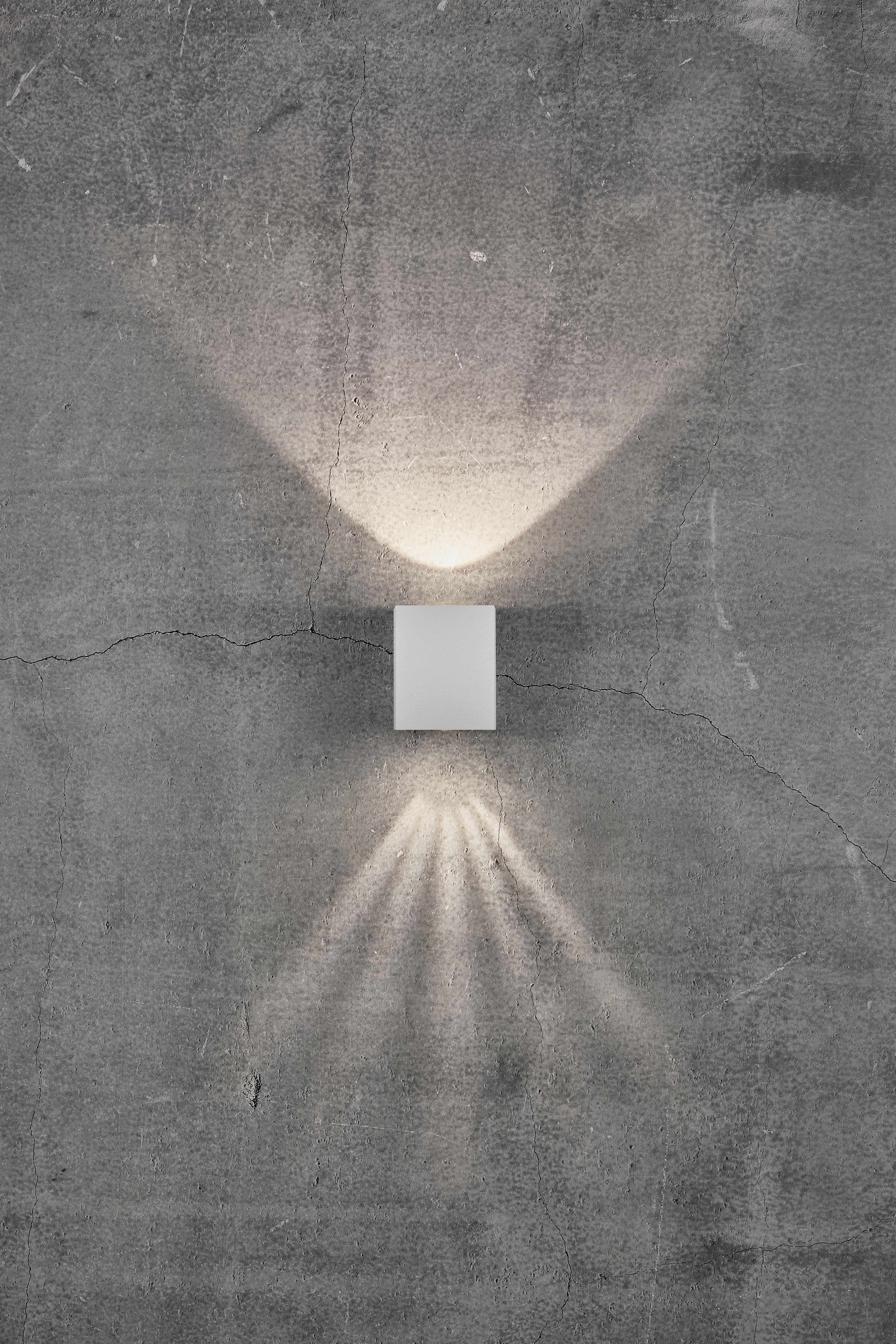 Kinkiet na szarej ścianie betonowej, emitujący miękki, symetryczny wzór świetlny w górę i w dół.