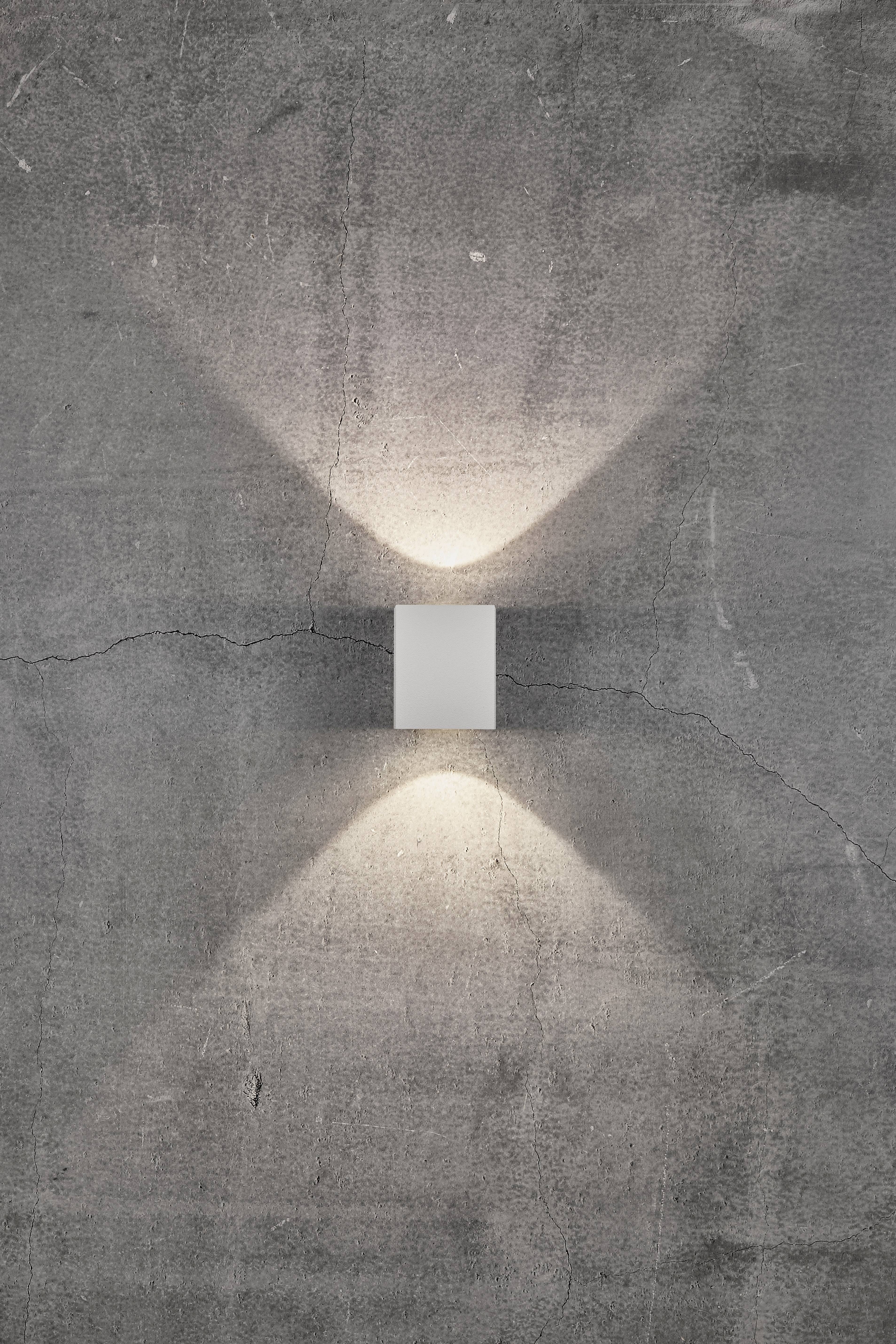 Kinkiet przy szarej ścianie betonowej, świecący w górę i w dół, tworzący dekoracyjne efekty świetlne.