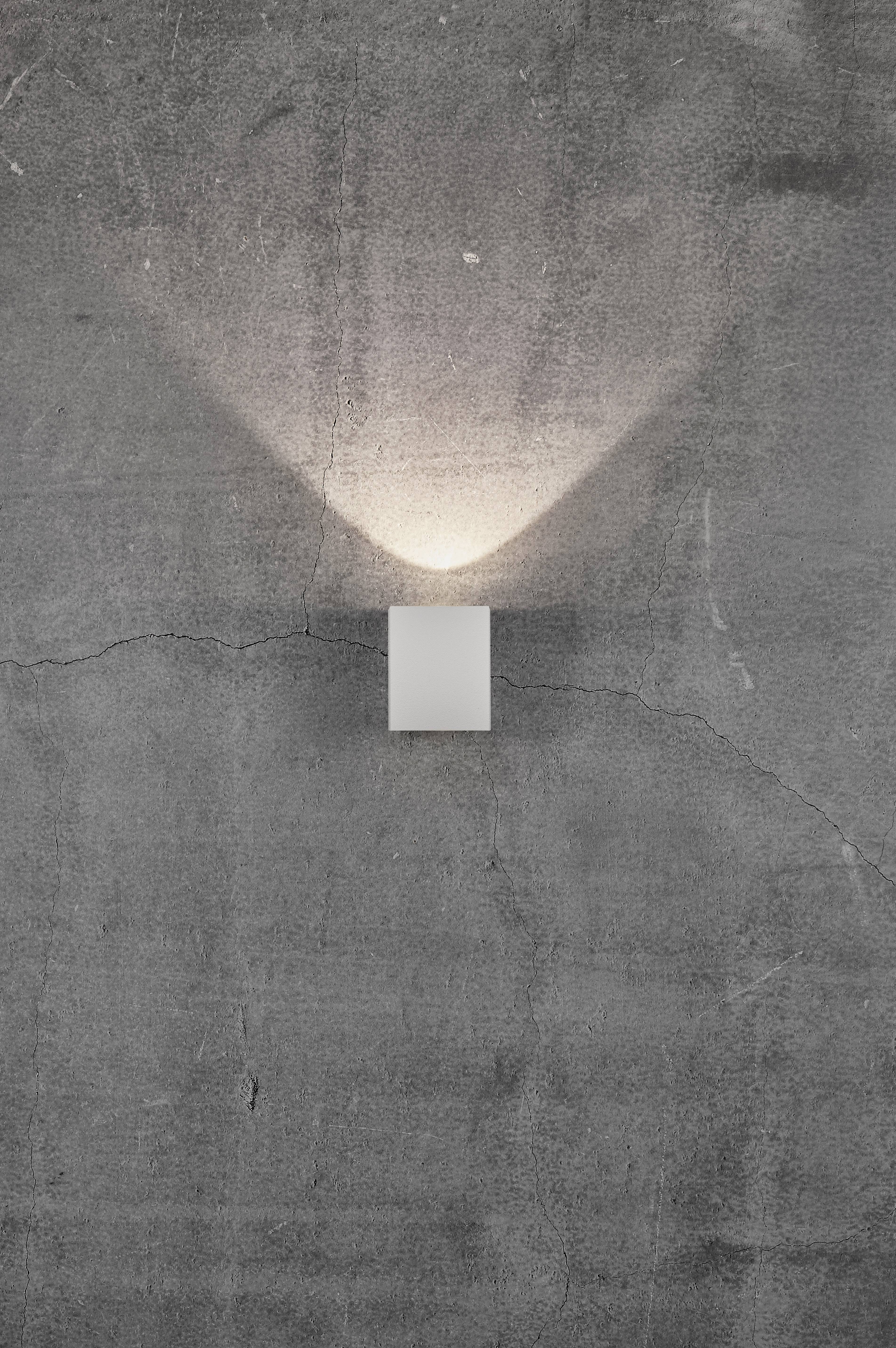 Kinkiet o nowoczesnym, minimalistycznym designie, rzucający ciepłe światło w kształcie litery V na szarą, lekko spękaną ścianę betonową.
