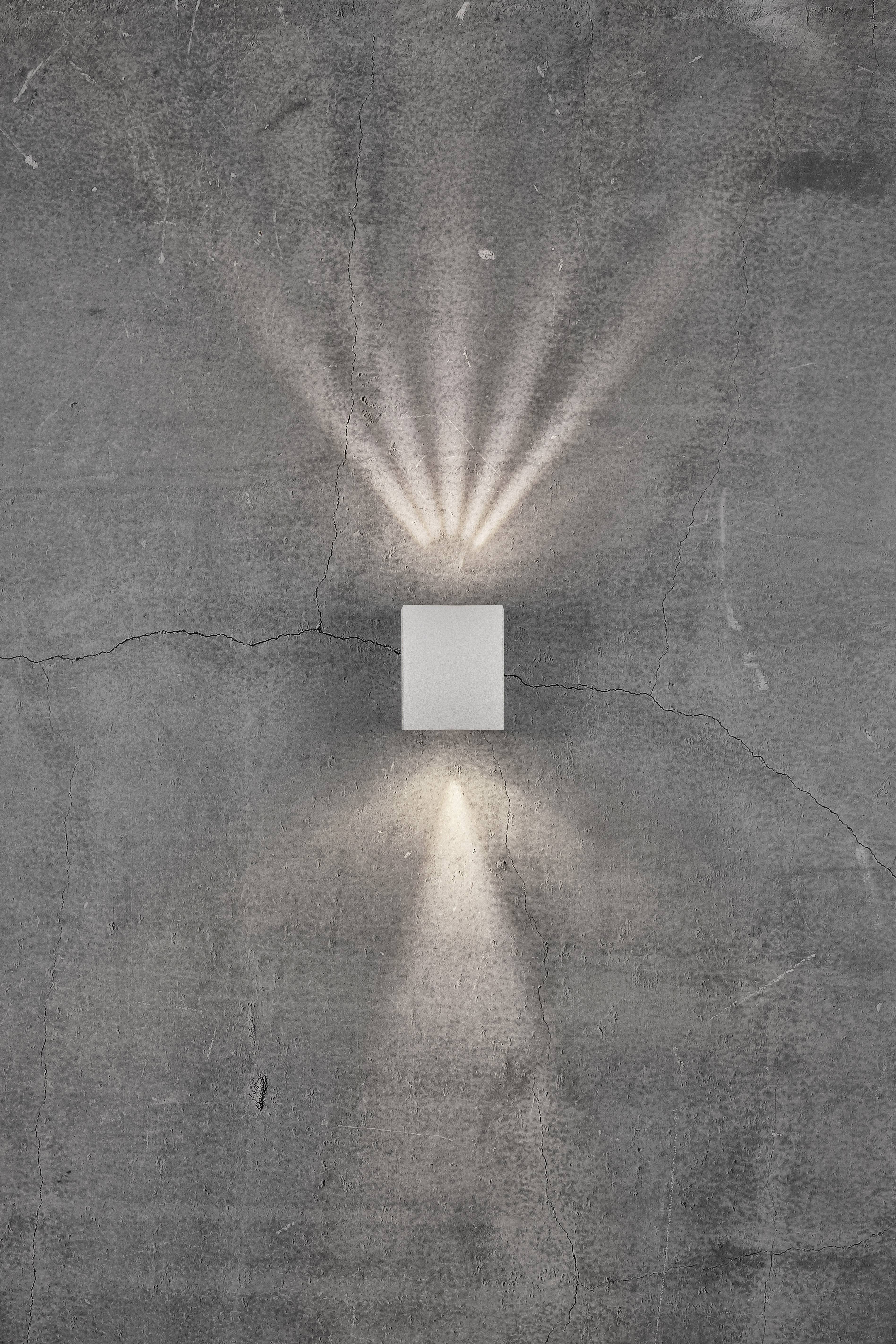 Kinkiet na betonowej ścianie, rzucający światło w górę i w dół w formie promieni.