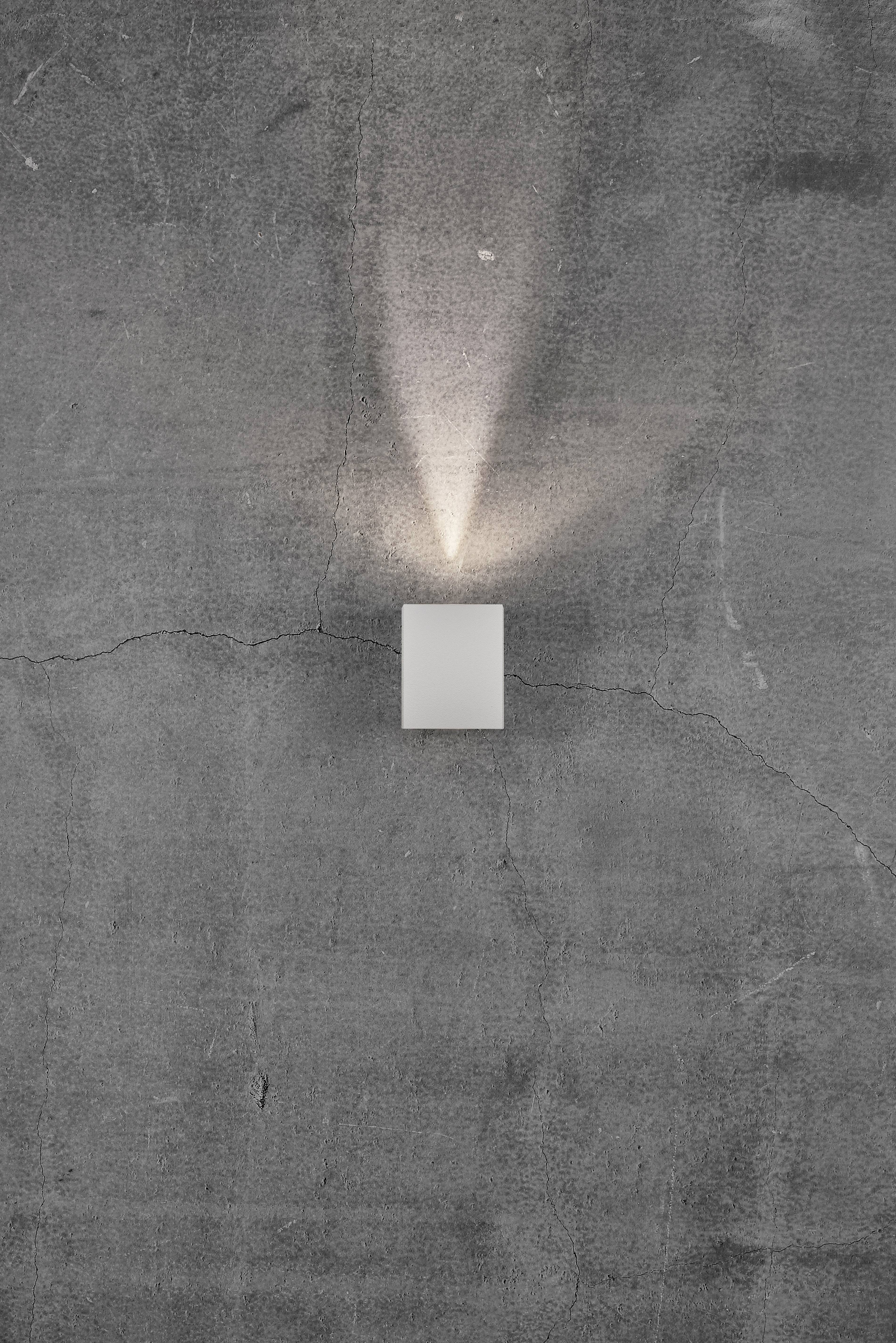 Kinkiet na szarym, popękanym tle betonowym, rzucający ciepłe światło skośnie w górę i w dół.