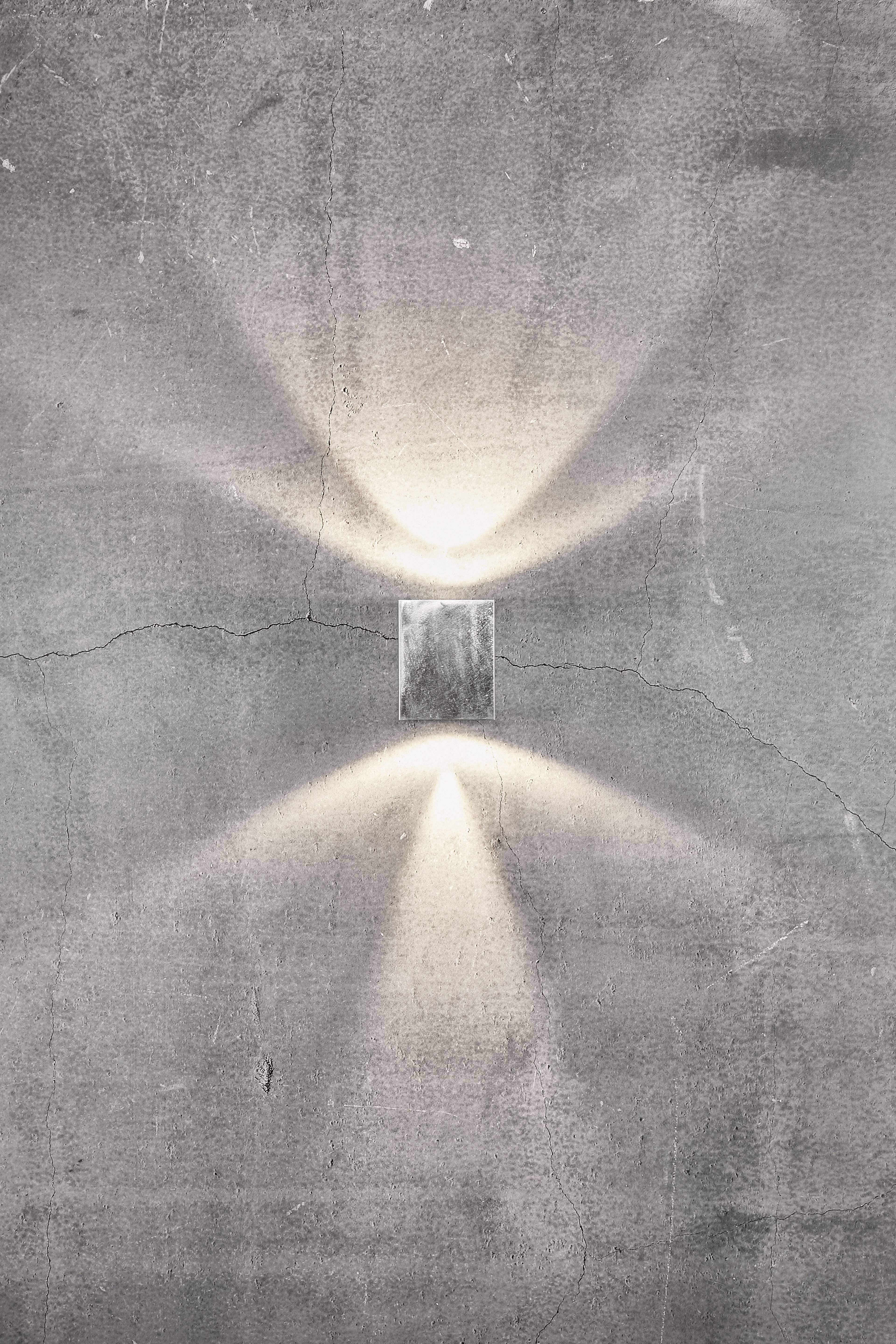 Kinkiet na szarej ścianie betonowej, rzucający ciepłe światło w górę i w dół, tworzący symetryczny wzór.