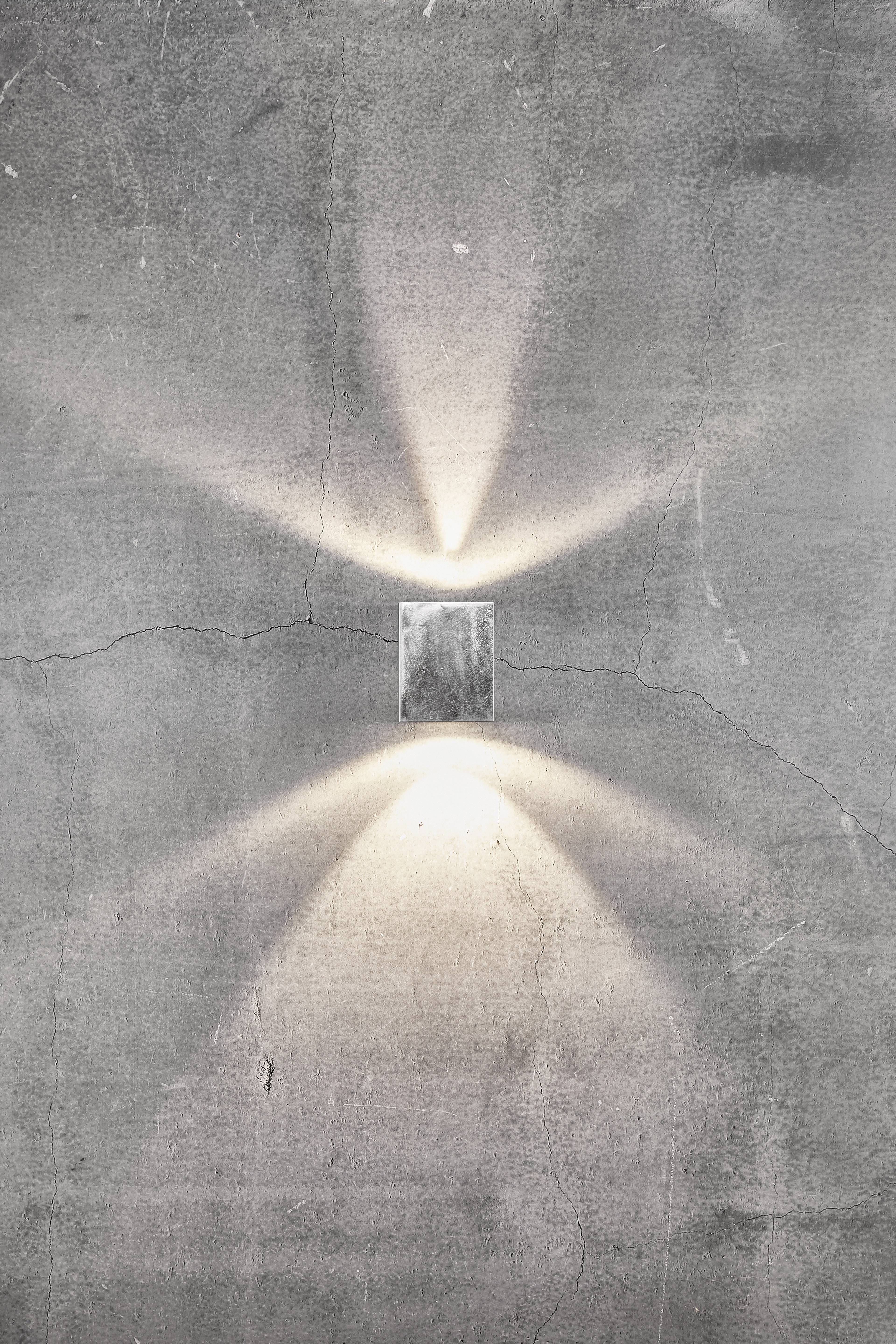 Kinkiet rzuca miękkie światło w górę i w dół na szarą betonową ścianę, tworząc dekoracyjny wzór świetlnych promieni.