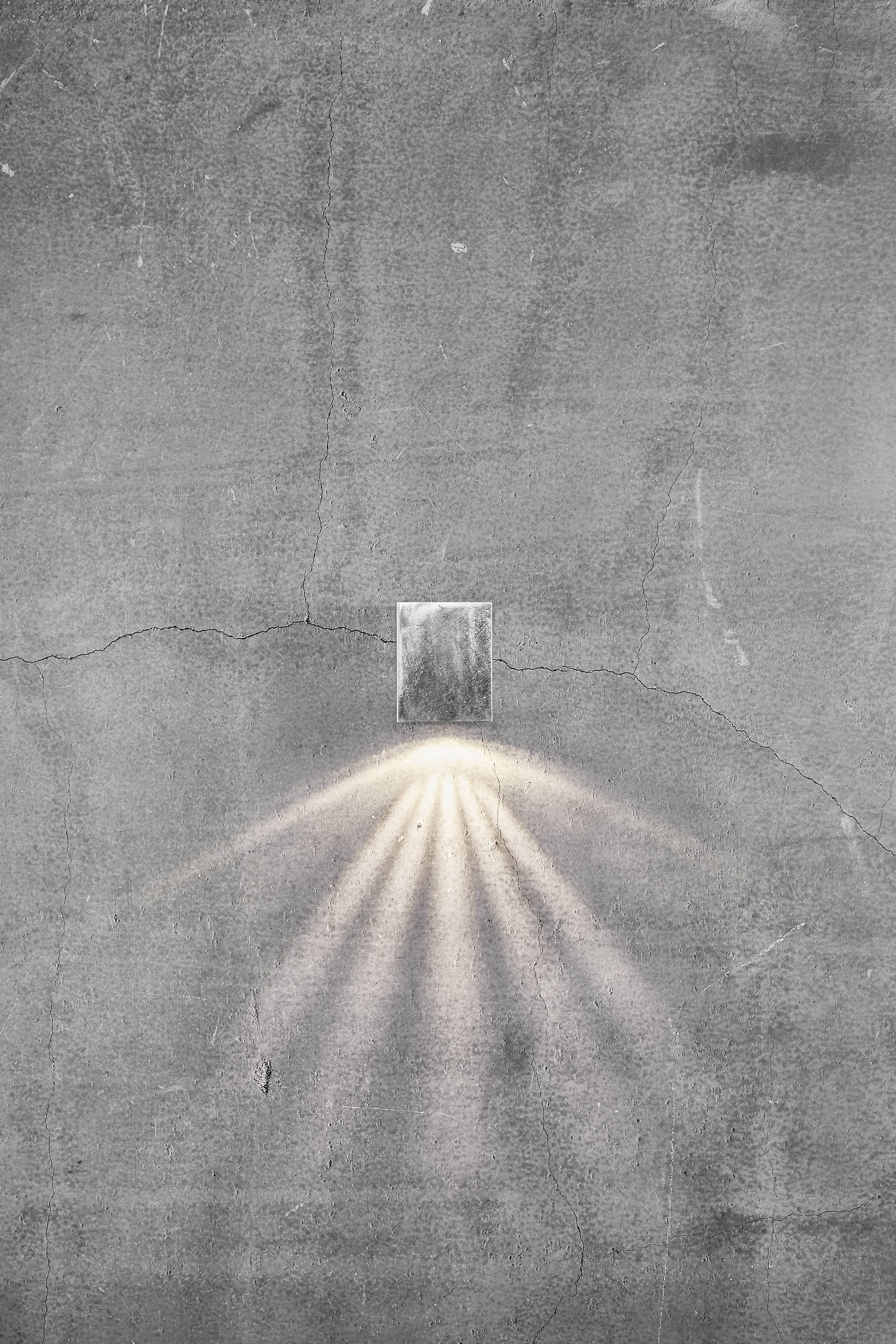 Wąski promień światła wpada przez małe okno w betonowej ścianie, tworząc wzory świetlne na szarej powierzchni.