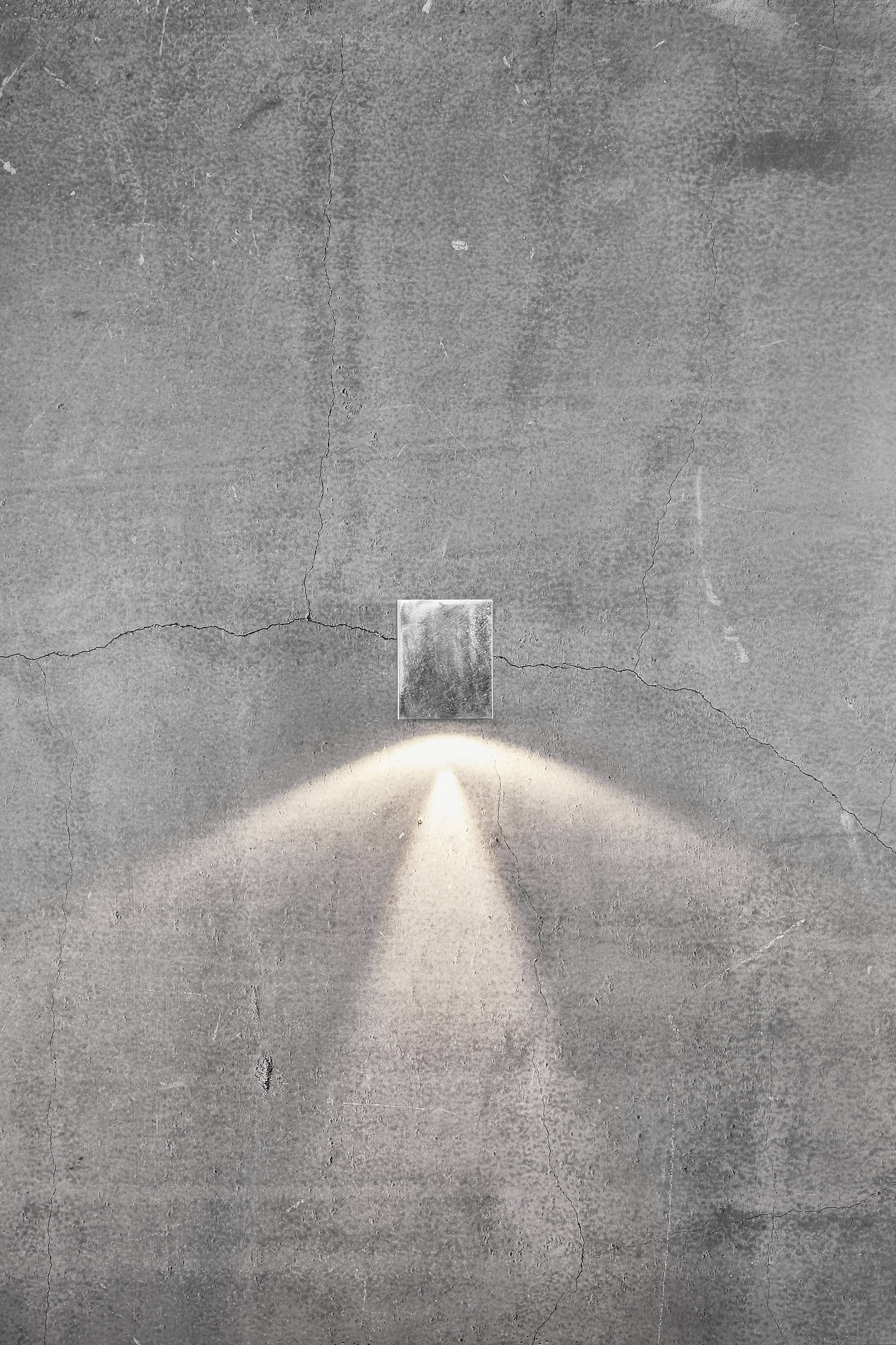 Pojedyncza, nowoczesna lampa ścienna rzuca ciepłe światło na szarą ścianę betonową, tworząc delikatny stożek świetlny skierowany w dół.