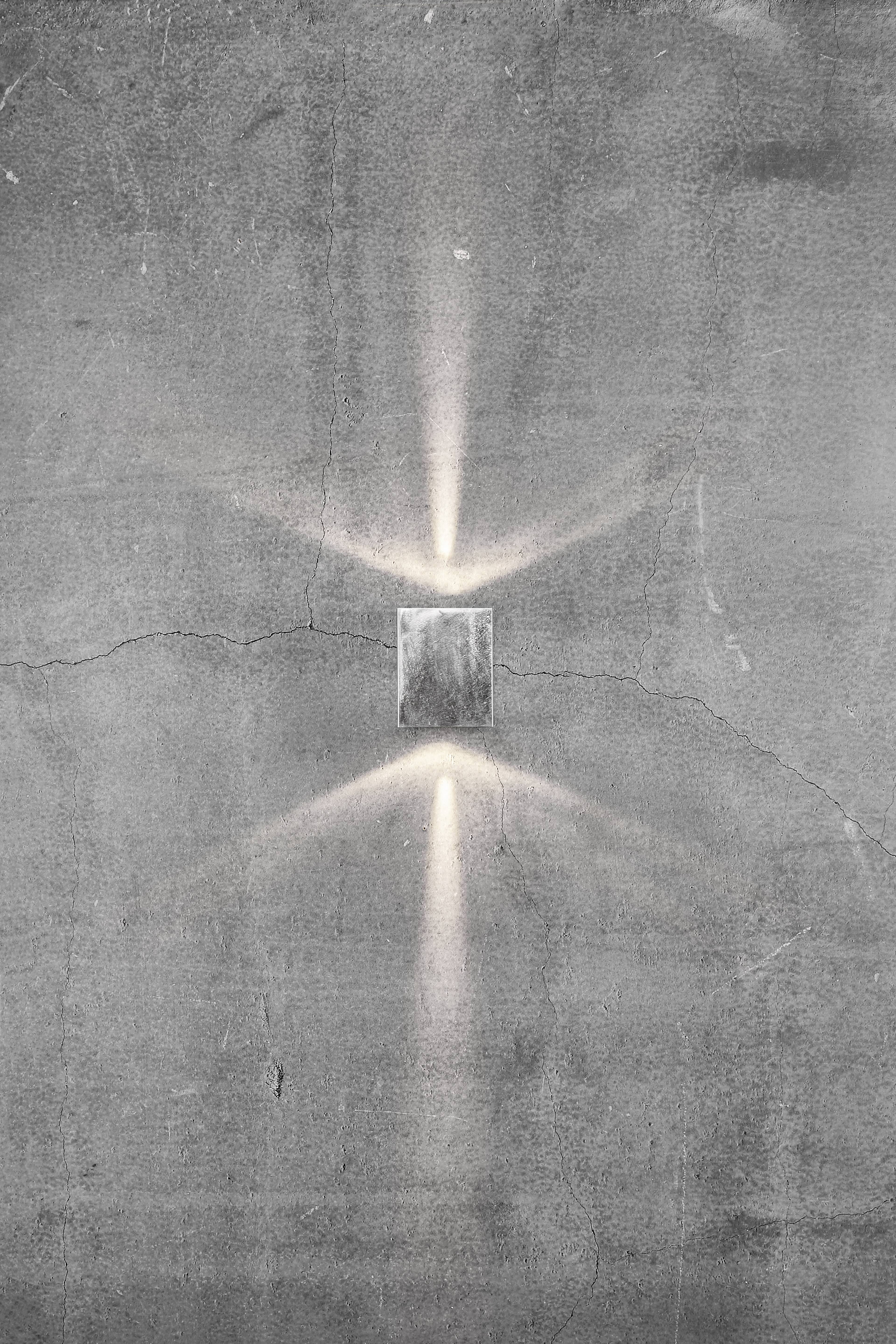 Prostokątna lampa ścienna, która emituje miękkie światło w górę i w dół, tworzy gwiazdkowaty wzór na szarej, teksturowanej ścianie.