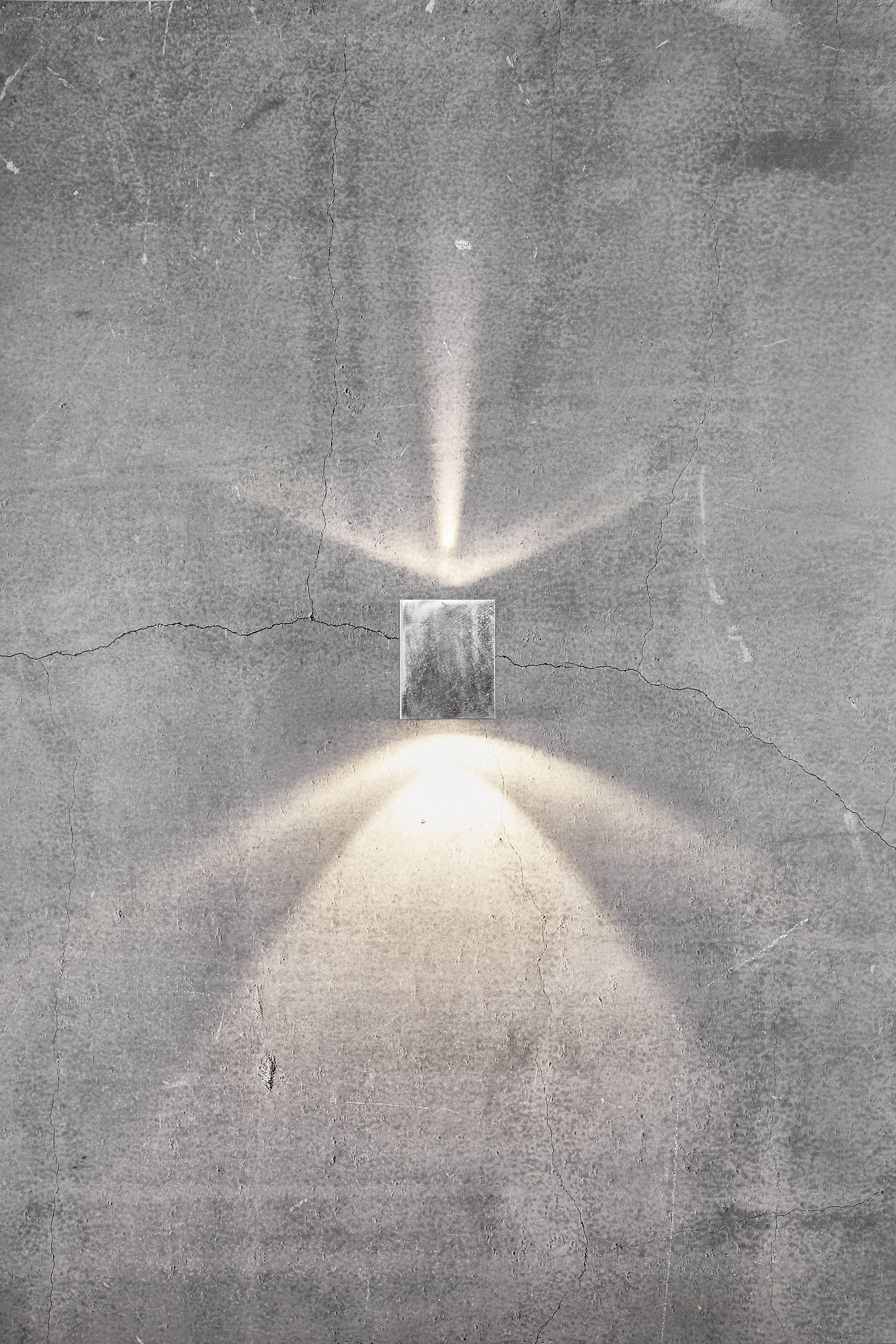 Lampa ścienna świeci na szarej, teksturowanej powierzchni, tworząc smugi światła rozchodzące się w górę i w dół.