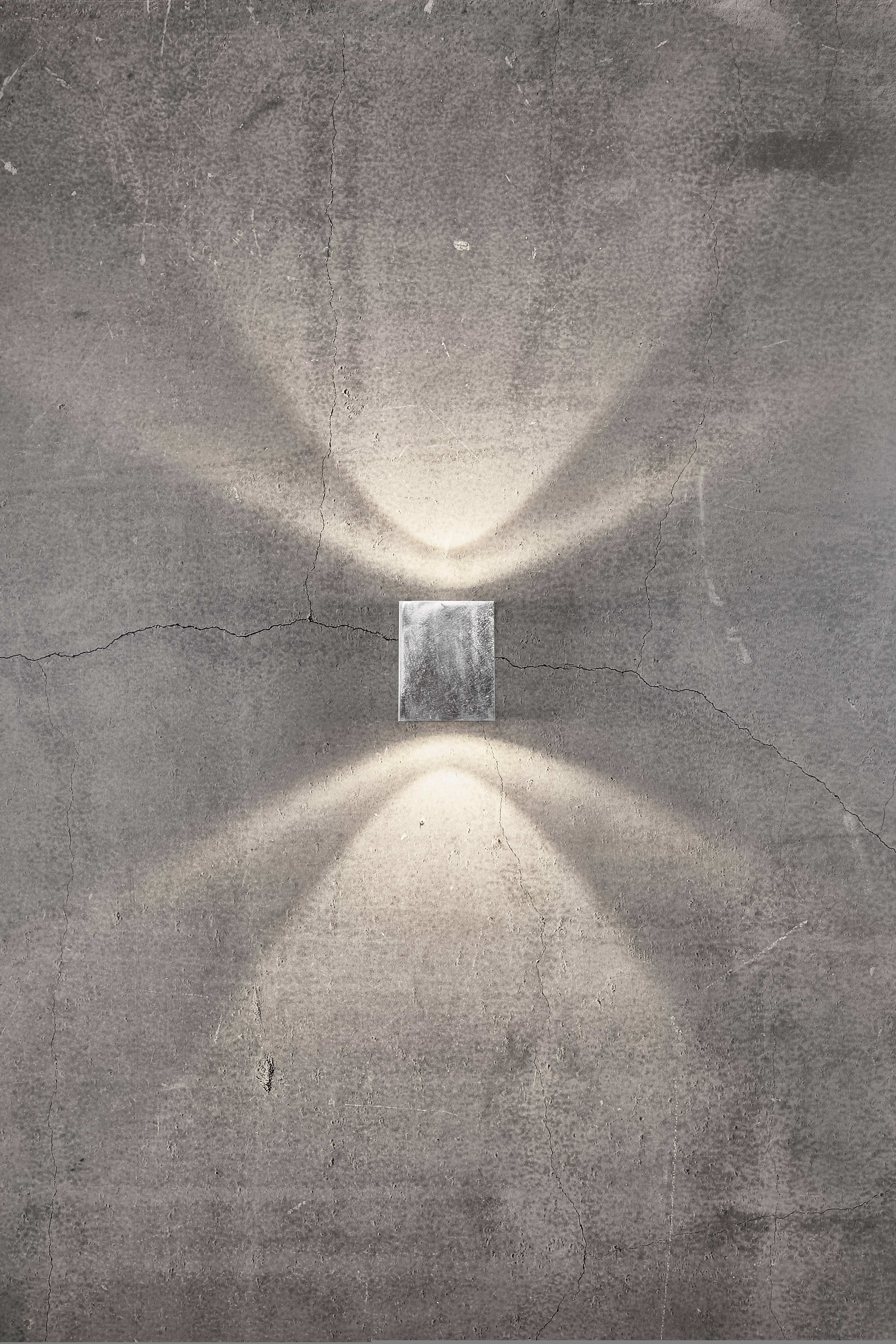 Szara lampa ścienna oświetla szorstką, spękaną ścianę betonową, rzucając światło w górę i w dół, tworząc nastrojowy wzór świetlny.