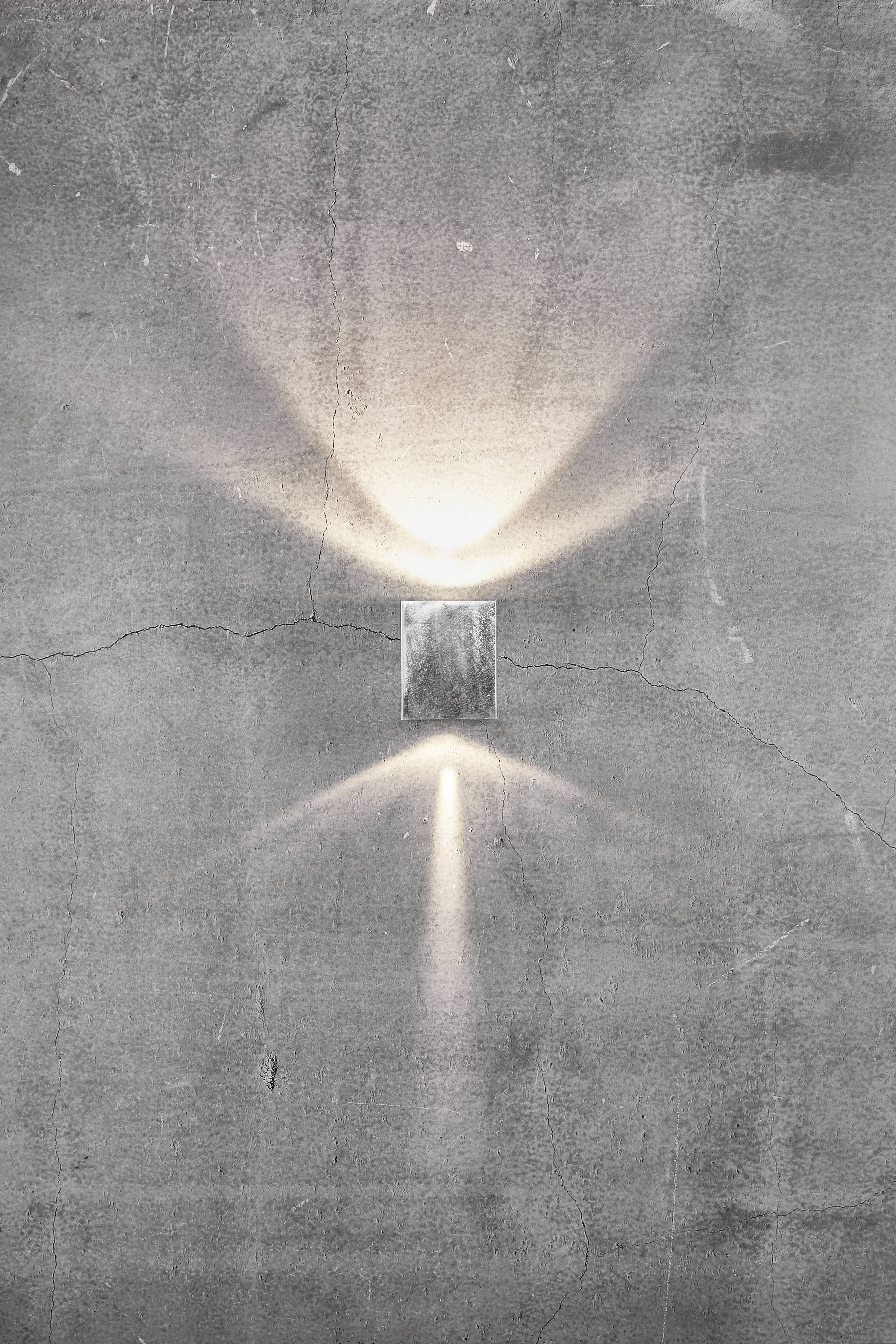 Lampa ścienna świeci na betonowej ścianie, tworząc symetryczne wzory świetlne skierowane w górę i w dół, otoczone spękaniami.