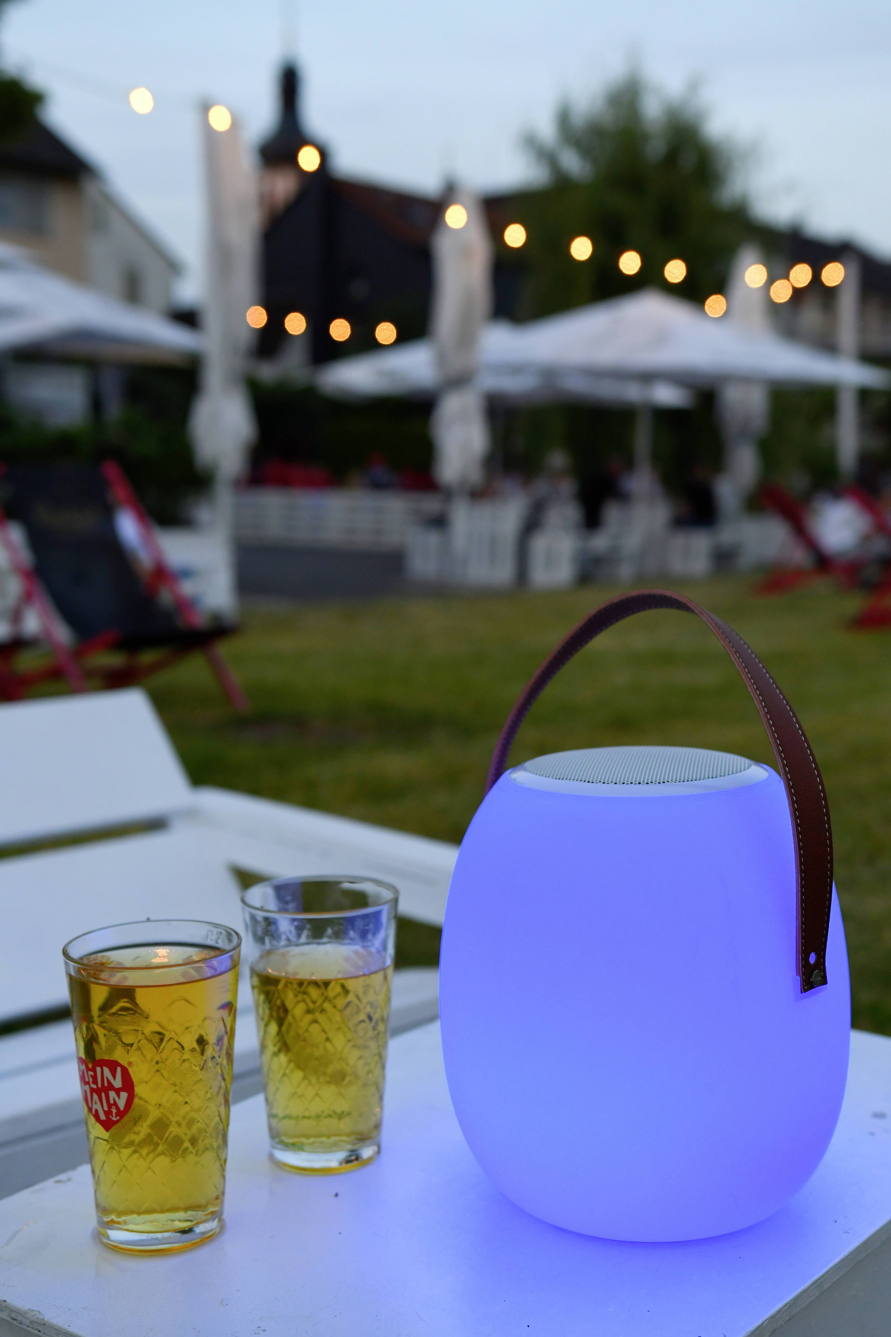 Dwa kieliszki z napojami stoją obok niebiesko świecącej lampy na stole na świeżym powietrzu, w tle widoczny jest girlandowy łańcuch świetlny.