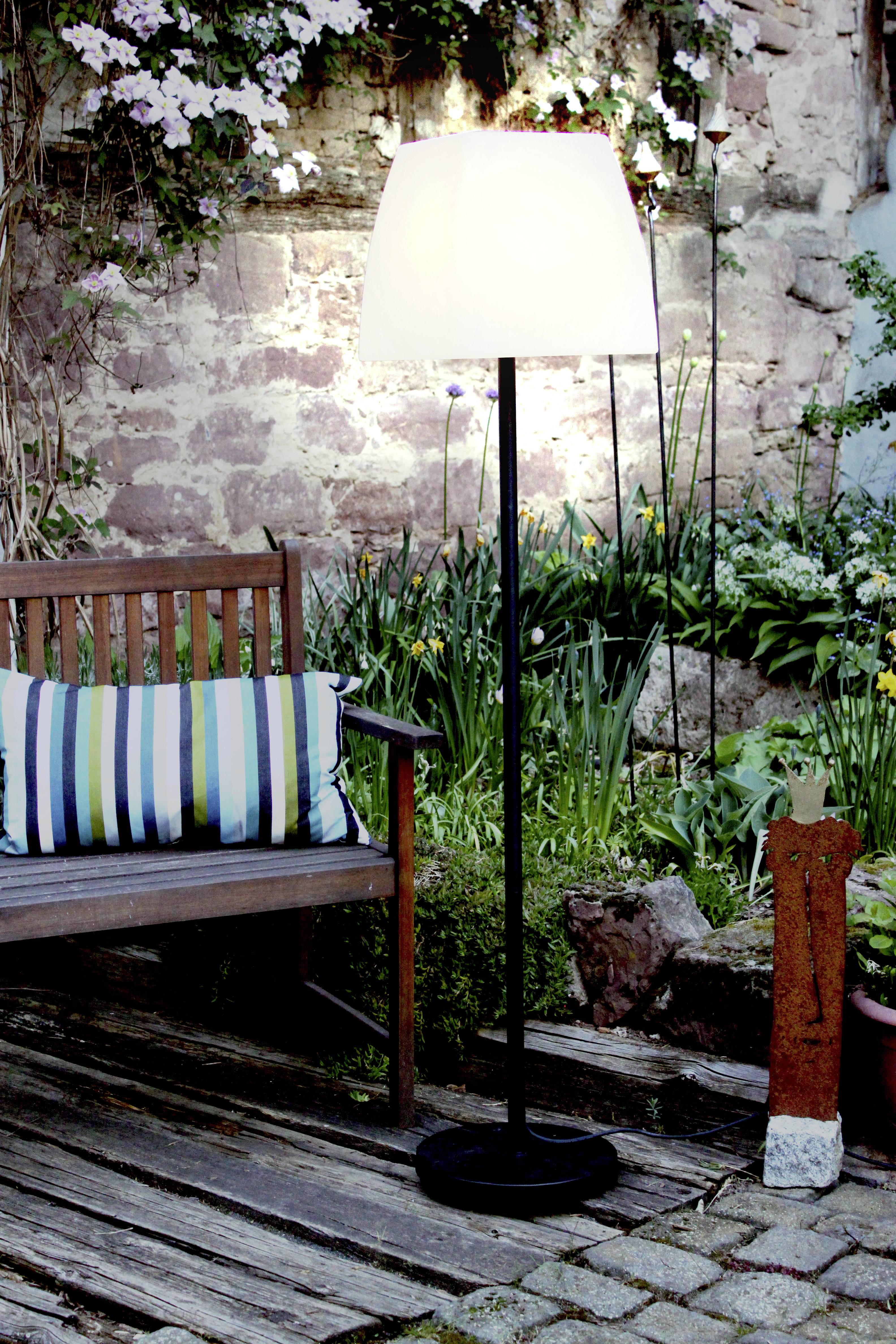 Lampa stojąca oświetla strefę wypoczynkową w ogrodzie, w skład której wchodzi ławka z poduszkami, otoczona kwiatami i roślinami, na tle starego muru kamiennego.