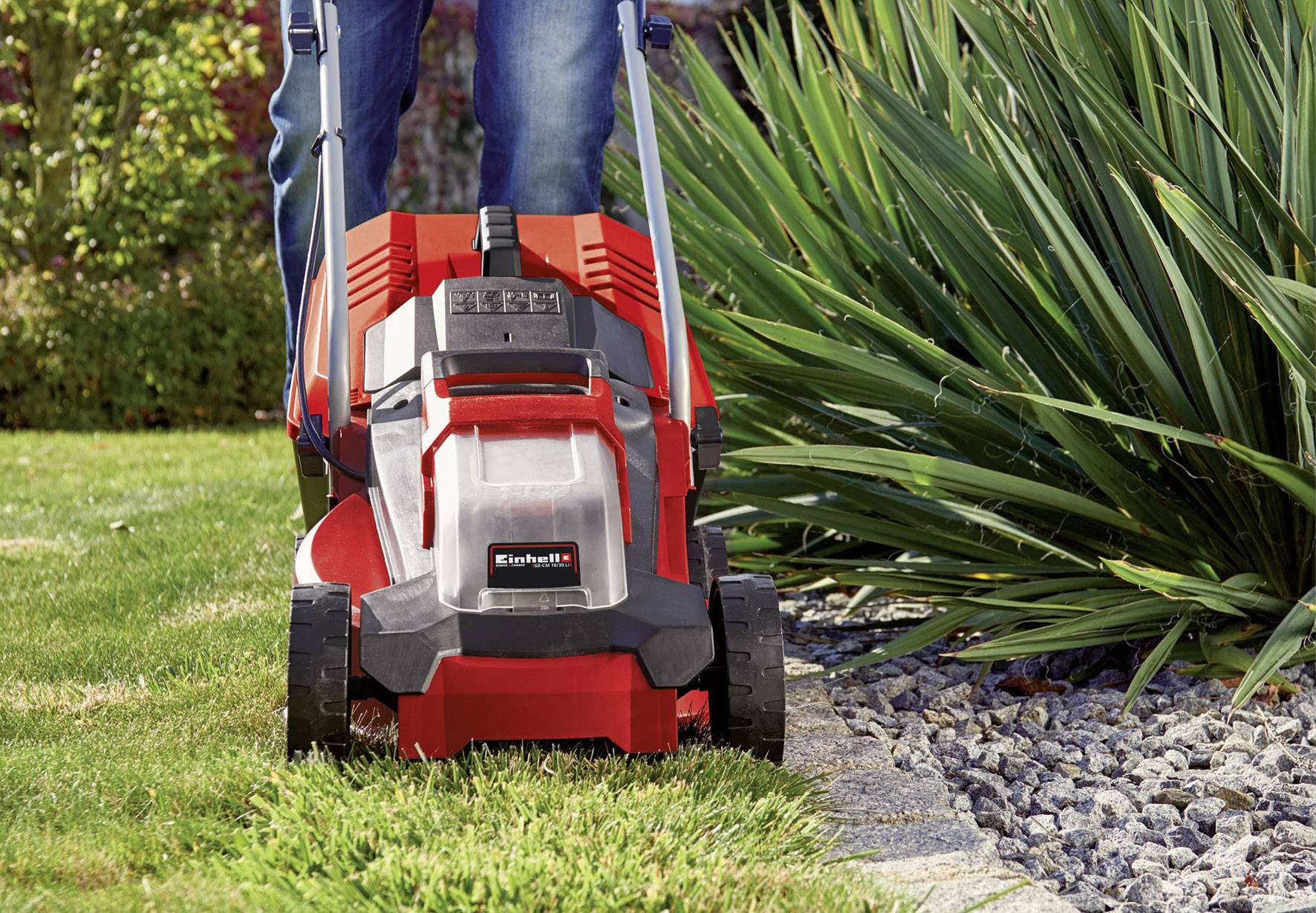 Osoba kosi trawę czerwoną kosiarką obok rabaty obramowanej kamieniami, pełnej zielonych roślin.