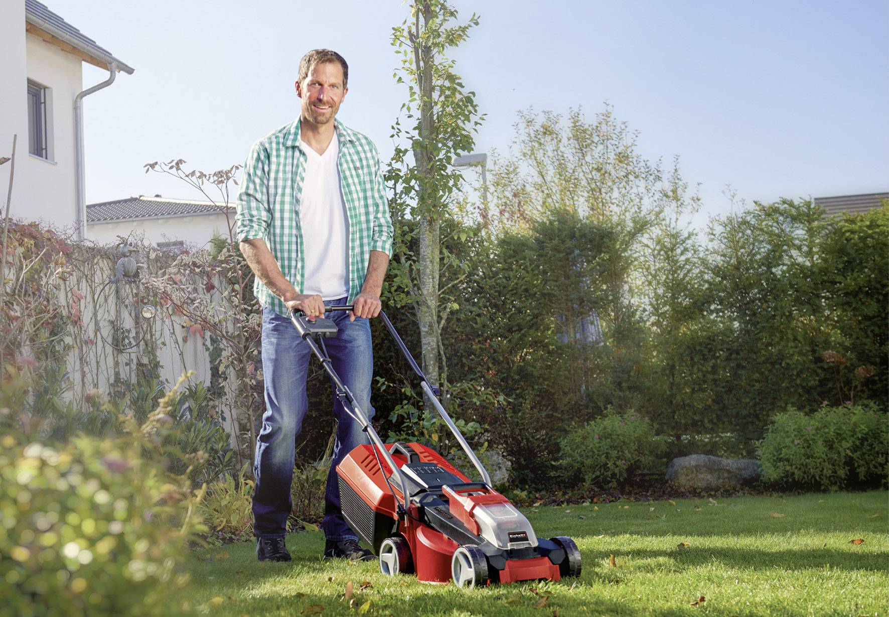 Mężczyzna uśmiechając się kosi trawę czerwonym kosiarką w słoneczny dzień w zielonym ogrodzie.