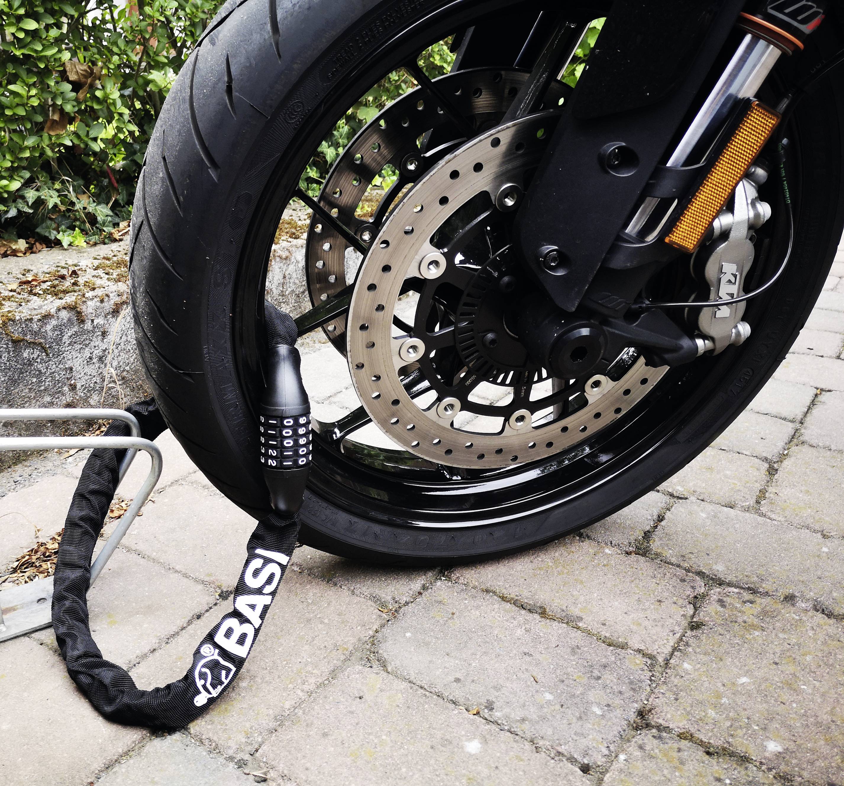 Motocykl stoi na wybrukowanej drodze. Przednie koło jest zabezpieczone ciężkim łańcuchem z kłódką.