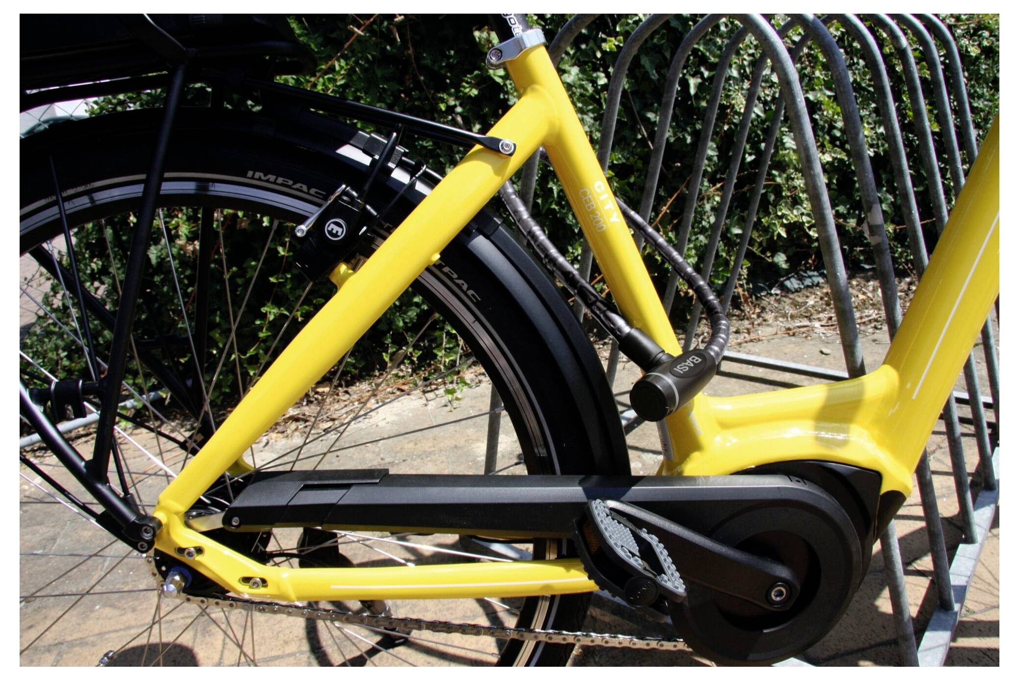 Żółta rama roweru przypięta do metalowego stojaka rowerowego, zbliżenie tylnego koła i łańcucha, z zielenią w tle.