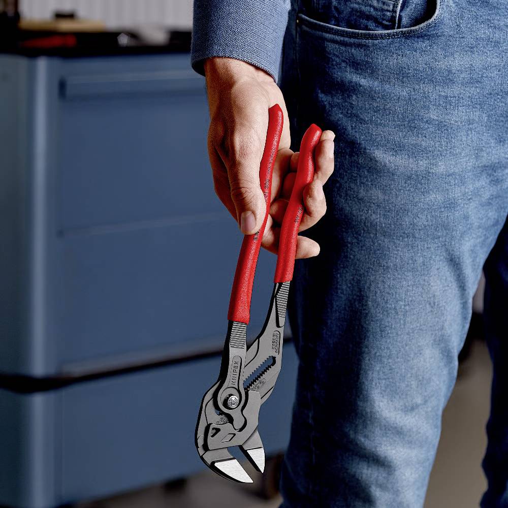 Osoba trzyma czerwone szczypce do rur w prawej ręce. W tle widać nieostro niebieski szafkę narzędziową.