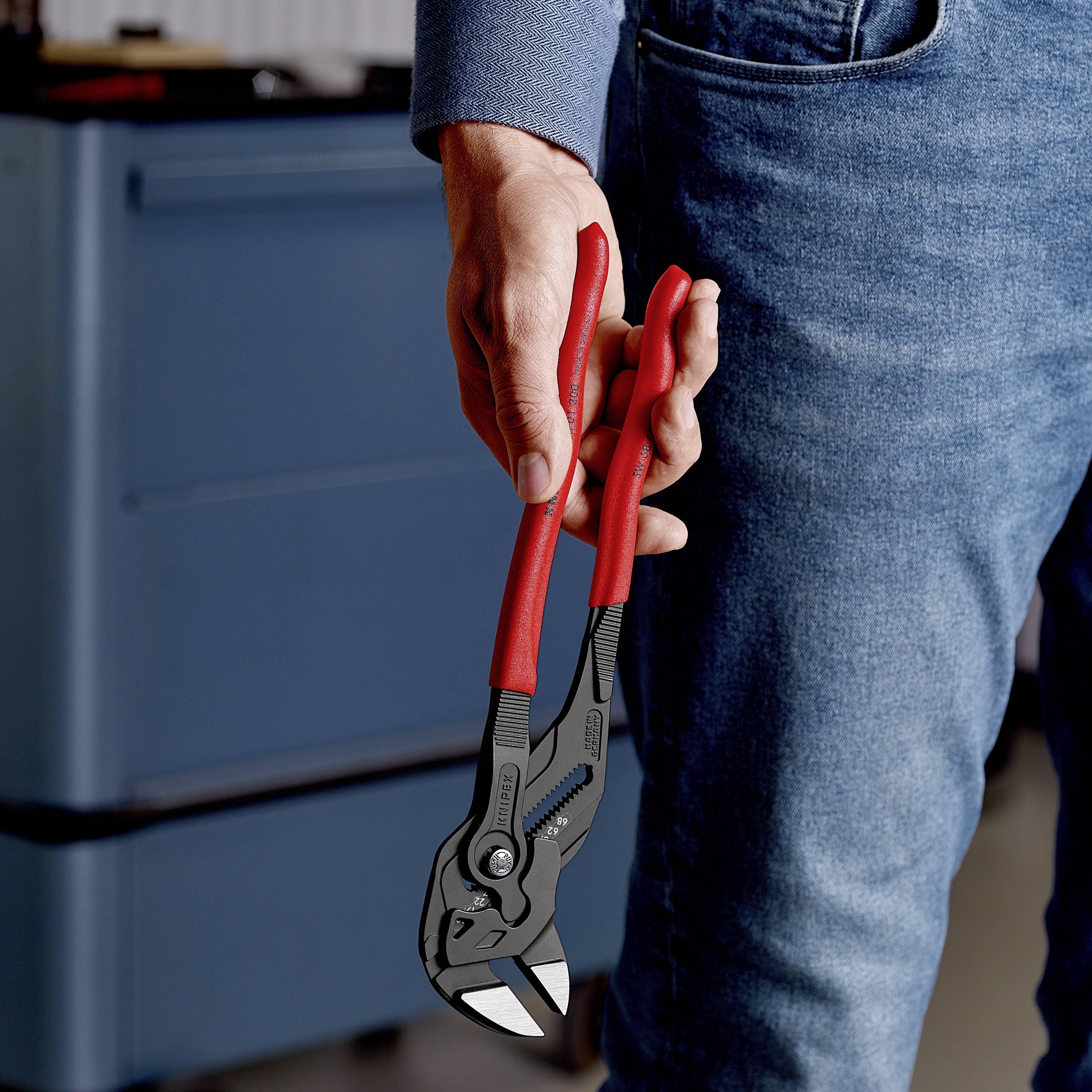 Dłoń trzyma czerwone szczypce blisko uchwytu w warsztacie, na tle niebieskich maszyn w tle.